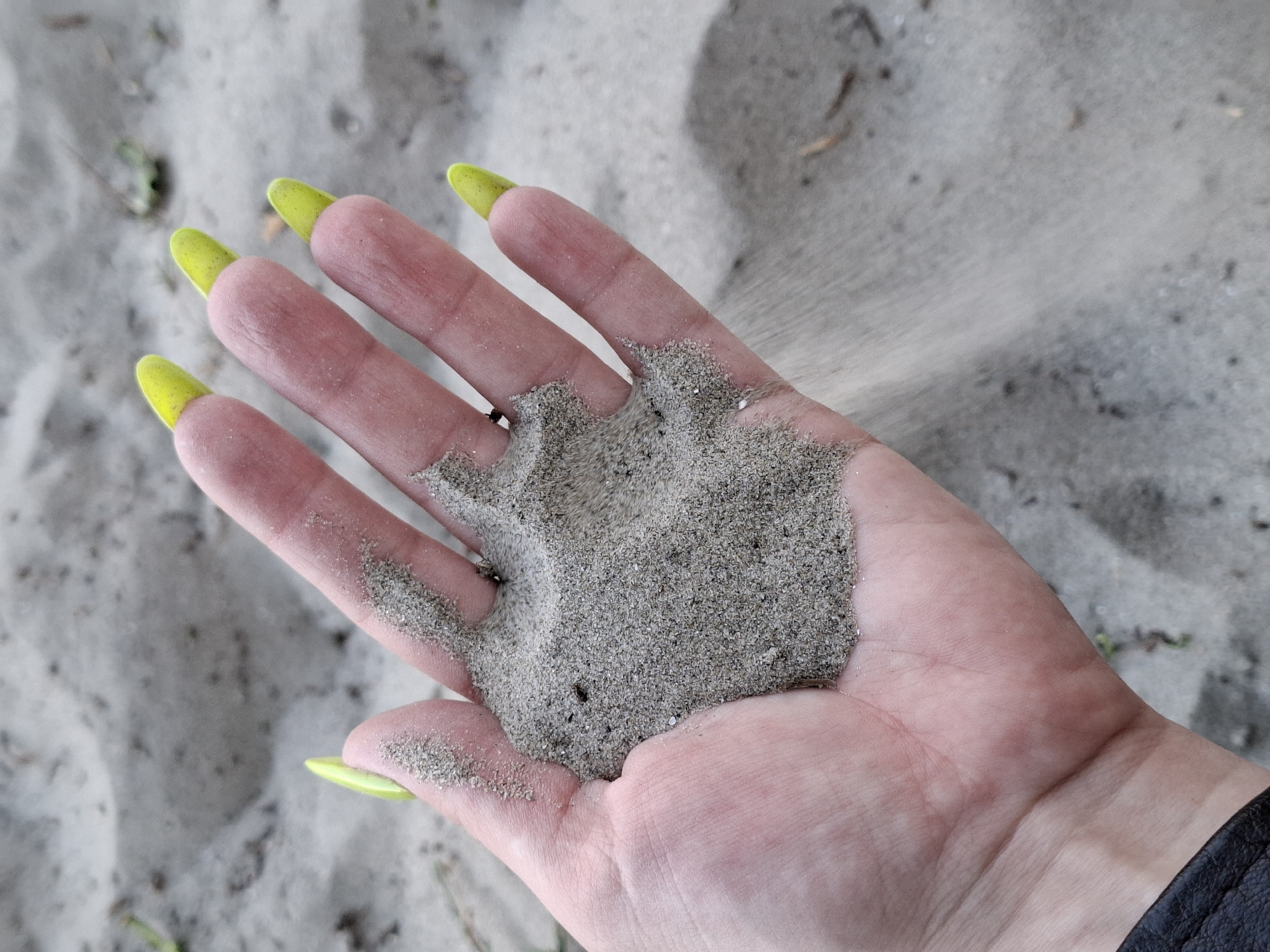 A human hand with neon green fingernails holds gray sand. The sand is partially falling between the fingers. The hand and sand are positioned against a backdrop of similar gray sand. A fragment of a black garment is visible in the lower right corner. Based on the sand characteristics, this image could be taken at Štrand beach in Novi Sad, Serbia.