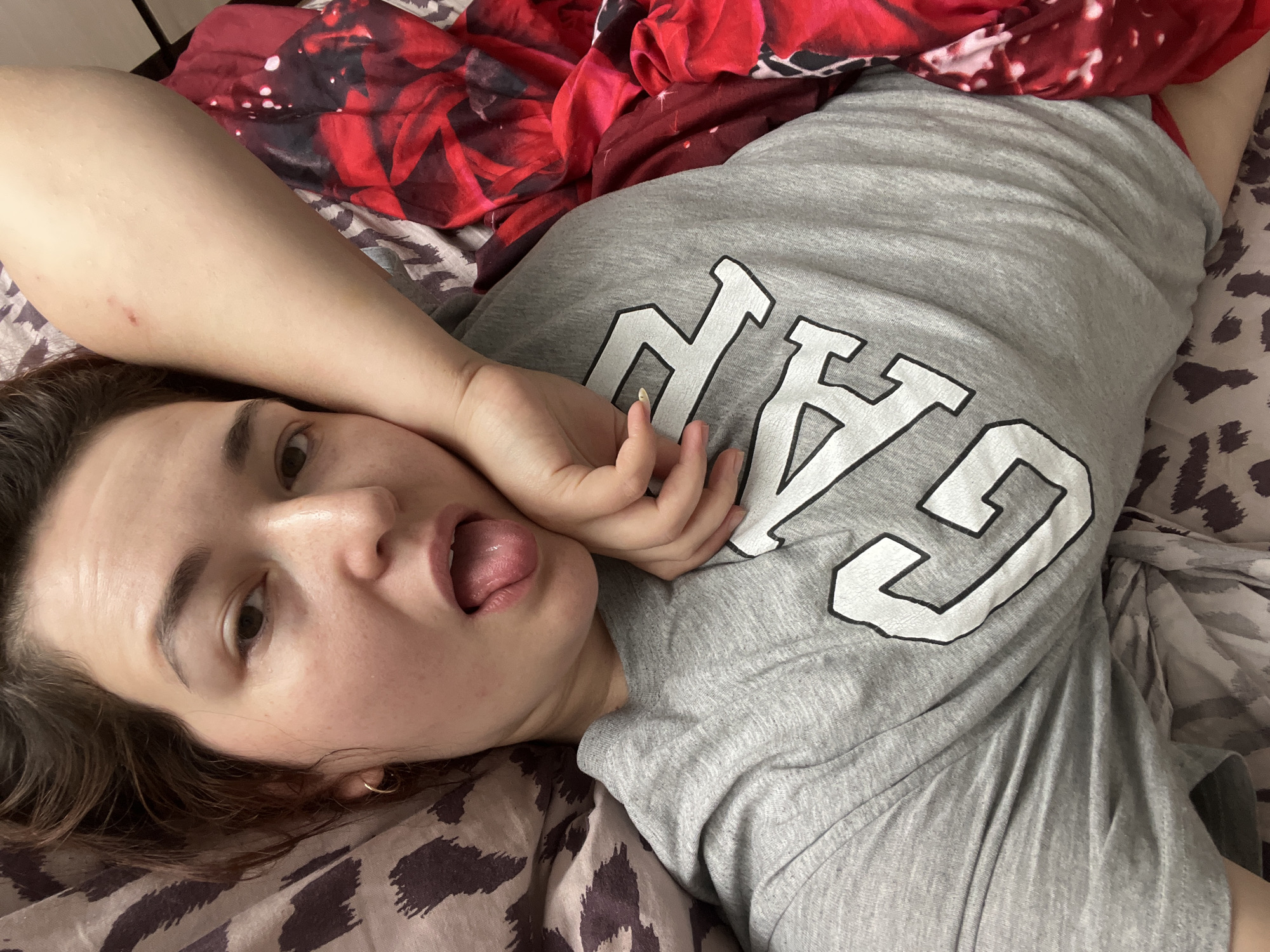 A young woman with dark hair is lying on a bed and sticking her tongue out at the camera. She is wearing a grey t-shirt with the word "GAP" printed on it in large white letters. Her right arm is bent, with her hand resting on her cheek. The bed is covered with patterned sheets, one with a leopard print design and the other with a vibrant red floral pattern. The image appears to have been taken indoors in a bedroom. There is no information available to determine the location in the city of Chelyabinsk, Russia.