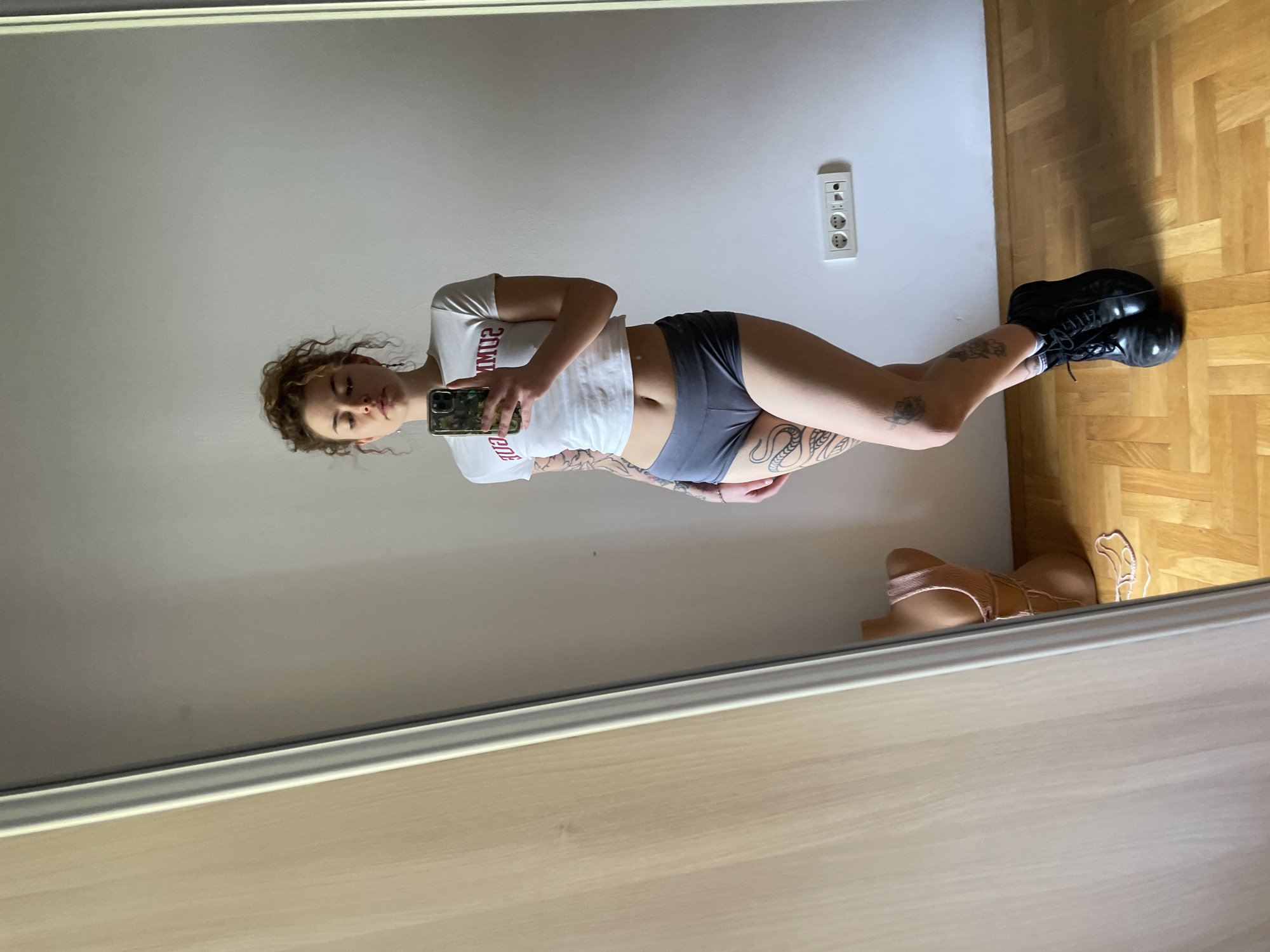 A woman is taking a selfie in front of a mirror. She is wearing a white t-shirt, gray shorts, and black boots. She has tattoos on her arms and legs. The background is a white wall with an electrical outlet and a wooden floor. The location is Boreti, Montenegro.