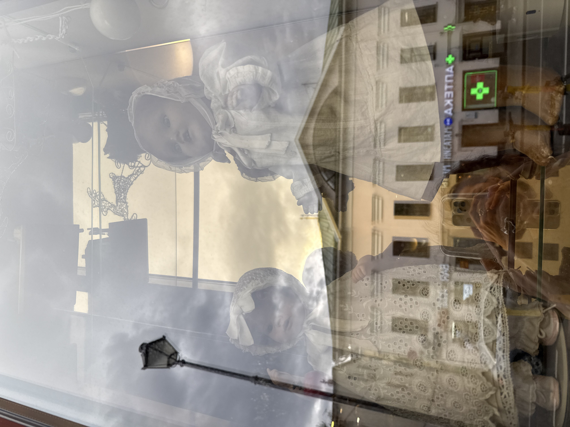 The image shows a reflection in a shop window of two antique dolls in white dresses. The doll on the right is larger and appears to be an adult doll, while the doll on the left is smaller, possibly a child's doll. Both dolls are wearing bonnets and intricate lace dresses. Behind the dolls, reflections show a street lamp, a decorative reindeer figure, and a white spherical object. Further in the background, the reflection of a building with a green roof is visible, along with signs for what appears to be a pharmacy, indicated by a green cross. The overall scene suggests a display in a shop window, with reflections from the street outside altering the view. The presence of the pharmacy sign and the Cyrillic text in the reflection suggests the location is likely in Moscow, Russia.