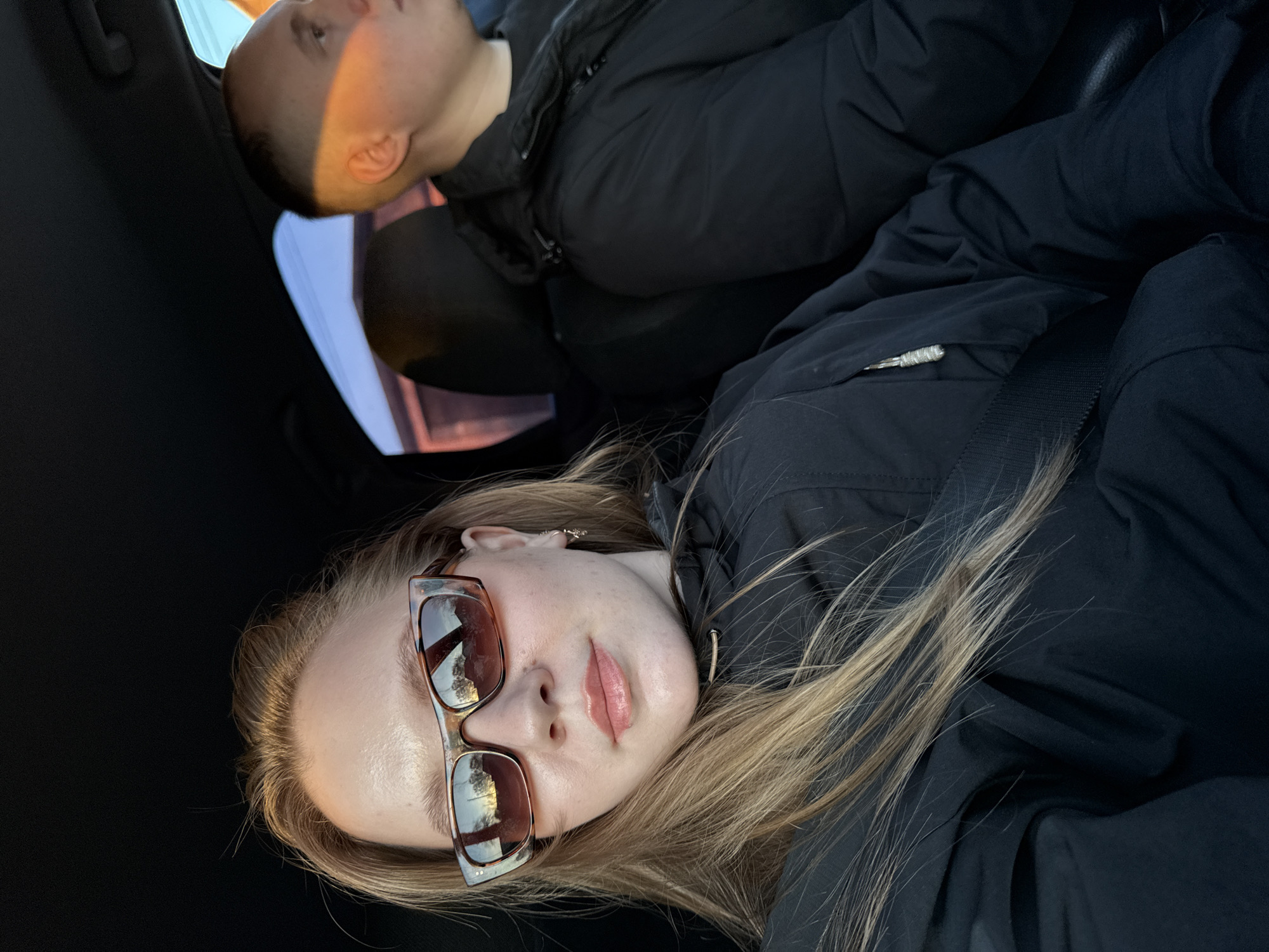 Two individuals are pictured inside what appears to be a car, likely captured during the "golden hour" of either sunrise or sunset, indicated by the warm, directional light.
In the foreground, a blonde woman is visible, wearing a dark jacket and stylish brown-tinted sunglasses, which reflect the external environment, possibly trees and sky. She has light makeup, including a rosy lip color, and looks directly towards the camera with a neutral to slight smile. Her long, blonde hair cascades over her shoulder and the dark fabric of her jacket. A seatbelt is visible across her chest.
Behind and above her, another individual with short, light hair is partially visible, wearing a black jacket. This person is looking away from the camera, seemingly towards the light source or resting, with a warm, orange glow illuminating the side of their face. Their head is tilted upwards.
The interior of the car is dark, creating a strong contrast with the exterior light filtering through the windows. The overall setting is intimate, capturing a moment within a vehicle. There is no visible text. The location context provided for this scene is Chaikovsky, Russia.