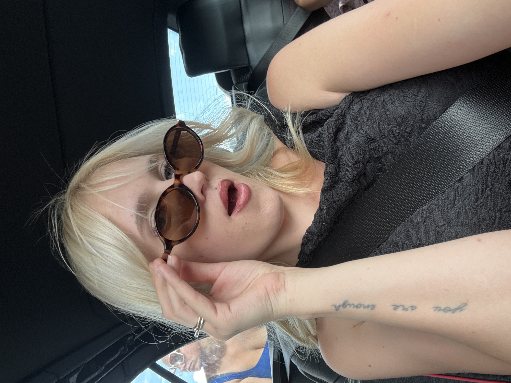 In an unknown city, a blonde woman with sunglasses and a black top is sitting in a car. She is holding her sunglasses with her right hand. Her mouth is slightly open, revealing her teeth and a lip piercing. A tattoo on her left arm reads "you are enough." In the background, a person wearing a blue top and sunglasses is visible through the car window. The car's interior, including a black seatbelt and headrest, is also visible. The lighting suggests it is daytime, with sunlight filtering through the windows.