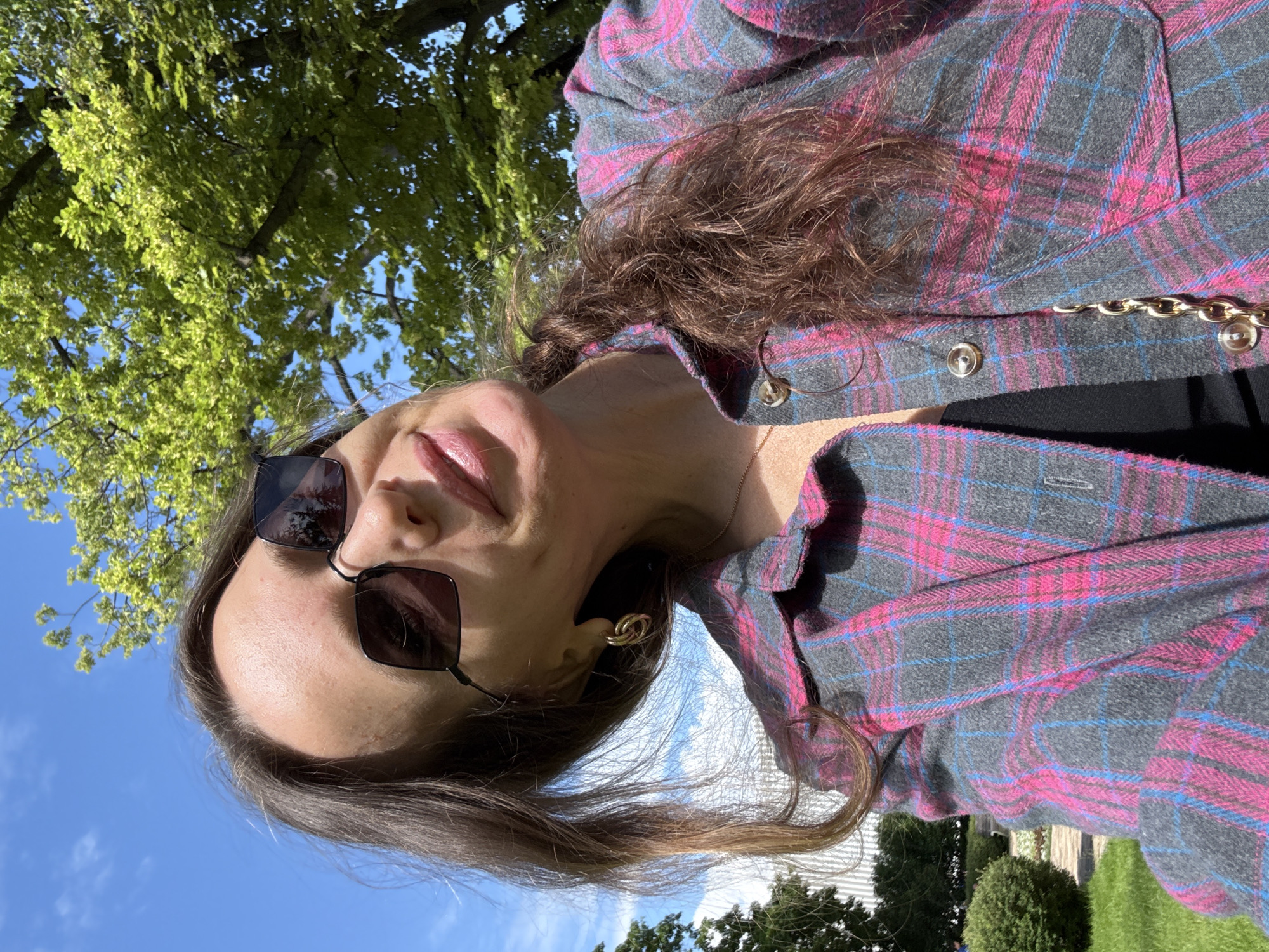 The image is a close-up, outdoor selfie of a woman. She is wearing a grey plaid shirt with pink and blue lines. She has on dark sunglasses with square frames and noticeable gold hoop earrings. Her hair is brown and appears to be styled in loose waves. The background shows a bright blue sky with some clouds and the green leaves of a tree. The sunlight suggests it is daytime, likely a clear and pleasant day. The woman is looking at the camera with a slight smile, and her lips are painted with pink lipstick. The overall mood of the image is casual and relaxed. There are no visible text elements in the image. The location context indicates this photo was taken in Moscow, Russia.
