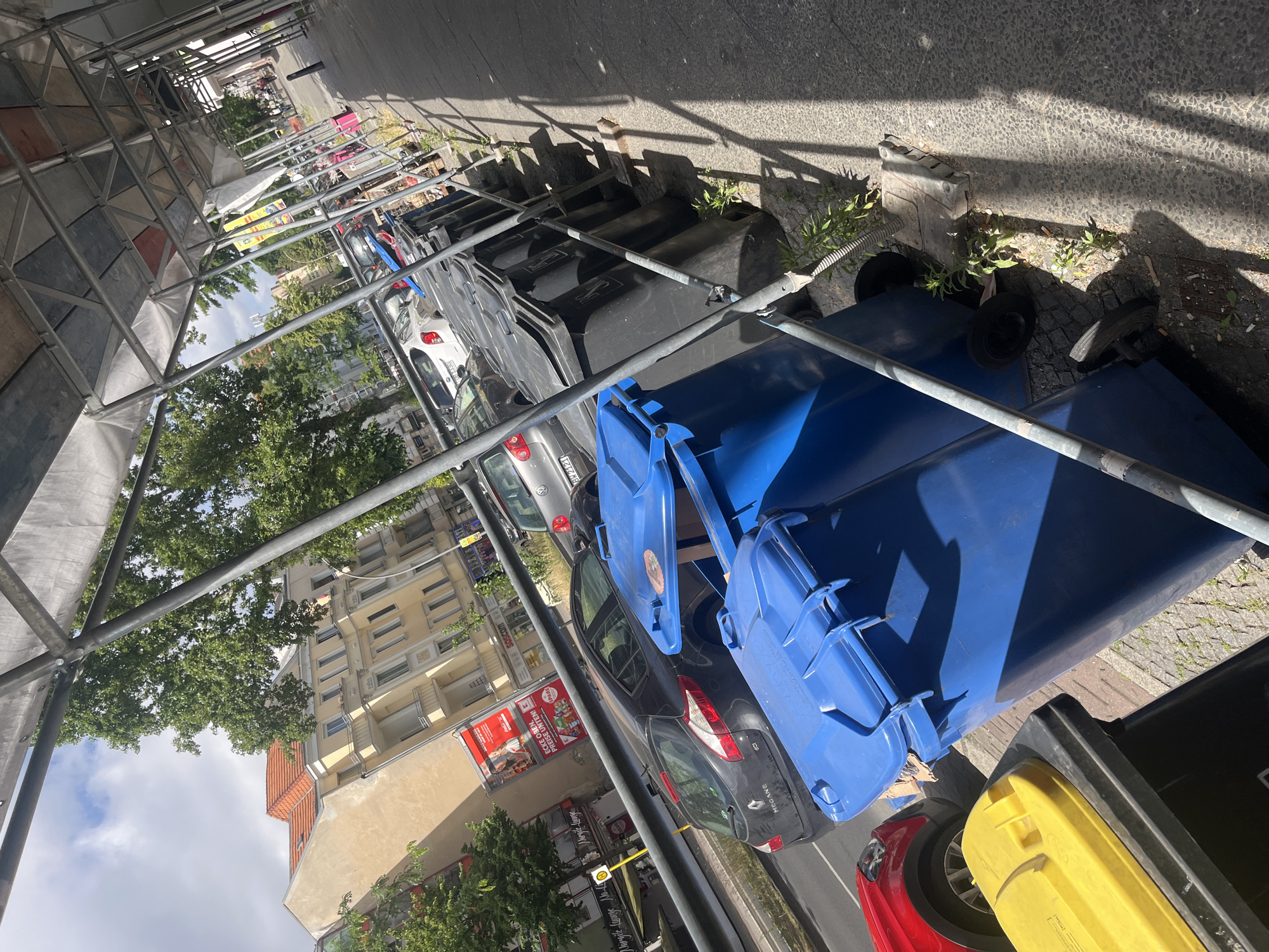 The image depicts a street scene in Berlin, Germany, characterized by temporary scaffolding along the sidewalk. In the foreground, several large trash bins are visible, including a bright blue one and a black one with a yellow lid. Behind these, a row of black trash bins lines the street. Cars are parked on the street, including a dark grey Renault Megane and a white Volkswagen Golf. Buildings with light-colored facades and red tile roofs are present in the background, along with mature trees. The sky is partly cloudy. The scene suggests an urban environment potentially undergoing construction or renovation due to the scaffolding.
