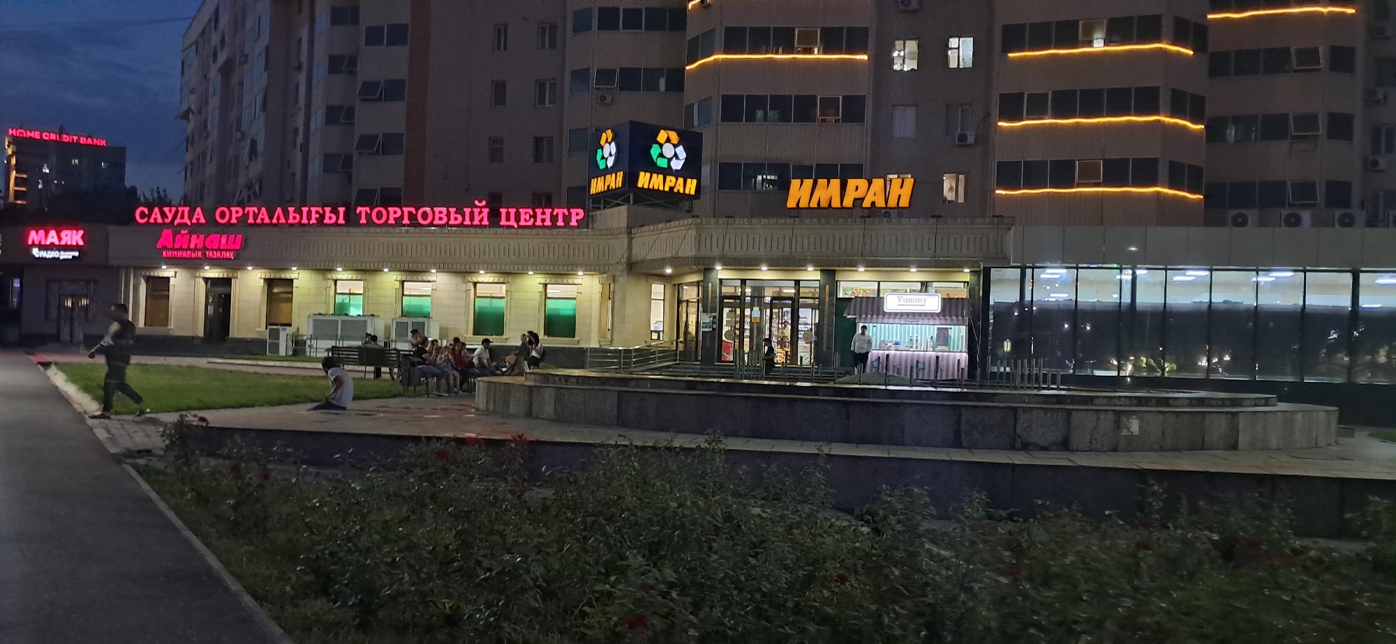 This media file shows a street-level view of a commercial building at dusk. The building has multiple businesses with illuminated signs, including "HOME CREDIT BANK," "МАЯК," "Айнаш," and "ИМРАН." The overall sign on the building reads "САУДА ОРТАЛЫҒЫ ТОРГОВЫЙ ЦЕНТР," indicating it's a shopping center. The building's facade features a modern design with large windows and illuminated architectural lighting. In front of the building, there's a paved area with a low stone planter and some greenery. A few people are visible: one person walking on the sidewalk, a group sitting on benches, and another individual near a food stall labeled "Yummy." The time of day appears to be evening, as the sky is dark and the building lights are on. The weather is not explicitly indicated but appears calm. The scene suggests a typical urban commercial area in the evening, with some patrons present.