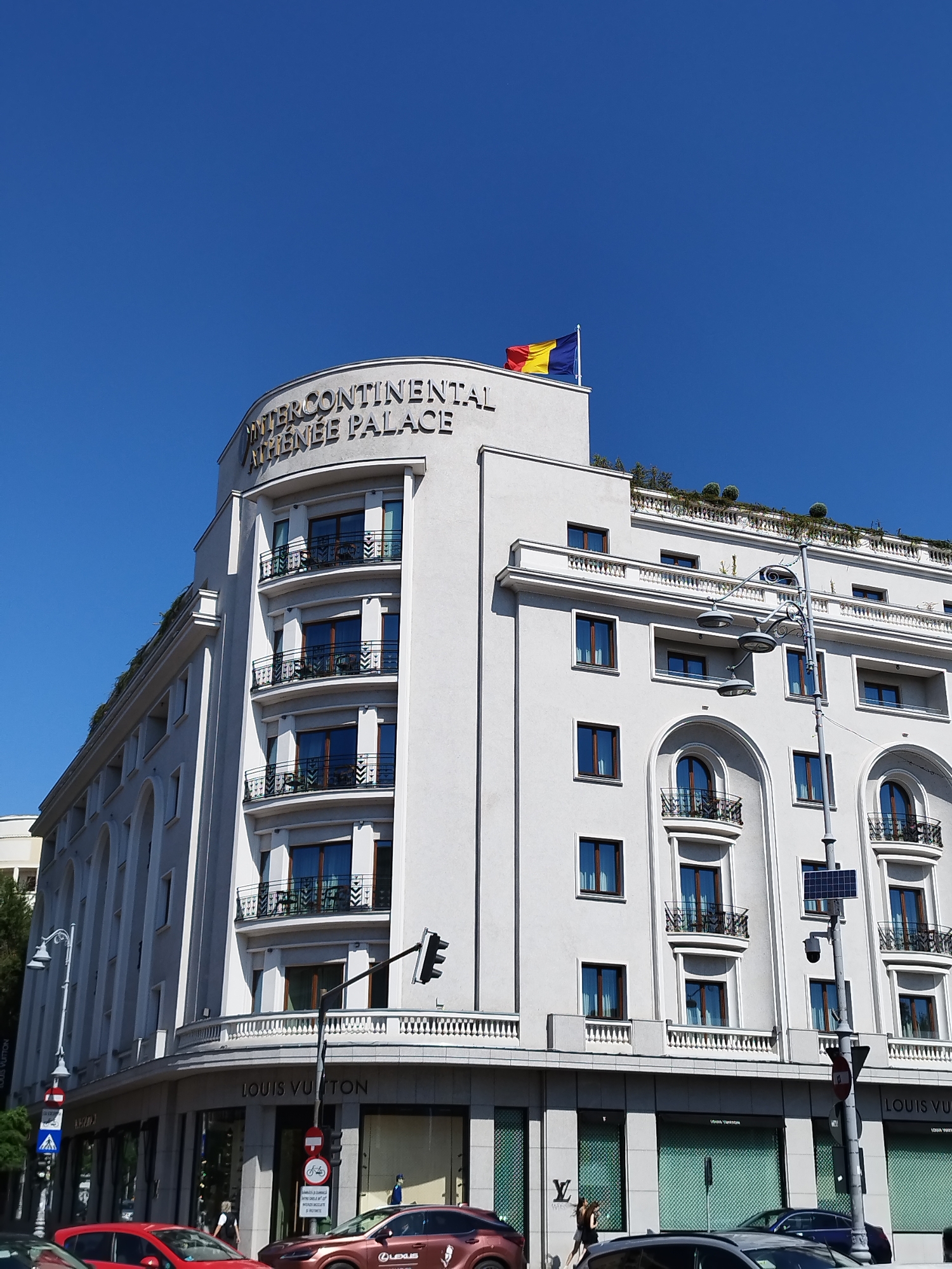 The image shows the InterContinental Atenee Palace hotel in Bucharest, Romania, under a clear blue sky. The hotel is a large, white, multi-story building with a distinctive curved corner. The name "INTERCONTINENTAL ATHENEE PALACE" is visible in dark lettering at the top of the curved section. A Romanian flag is flying from the top of the building.
The lower floors of the building house retail establishments, with "LOUIS VUITTON" signage prominently displayed. Several windows of the shops are visible, some with a distinctive green patterned grille.
In the foreground, a street scene is depicted with several cars. A red car is on the left, and a brown car with a "LEXUS" logo is on the right. Pedestrians are also visible on the sidewalk in front of the shops. A street light with multiple lamps and a traffic signal are present, along with various traffic signs. Trees and greenery are visible along the side of the building and on the rooftop terrace. The architecture suggests a European city setting.