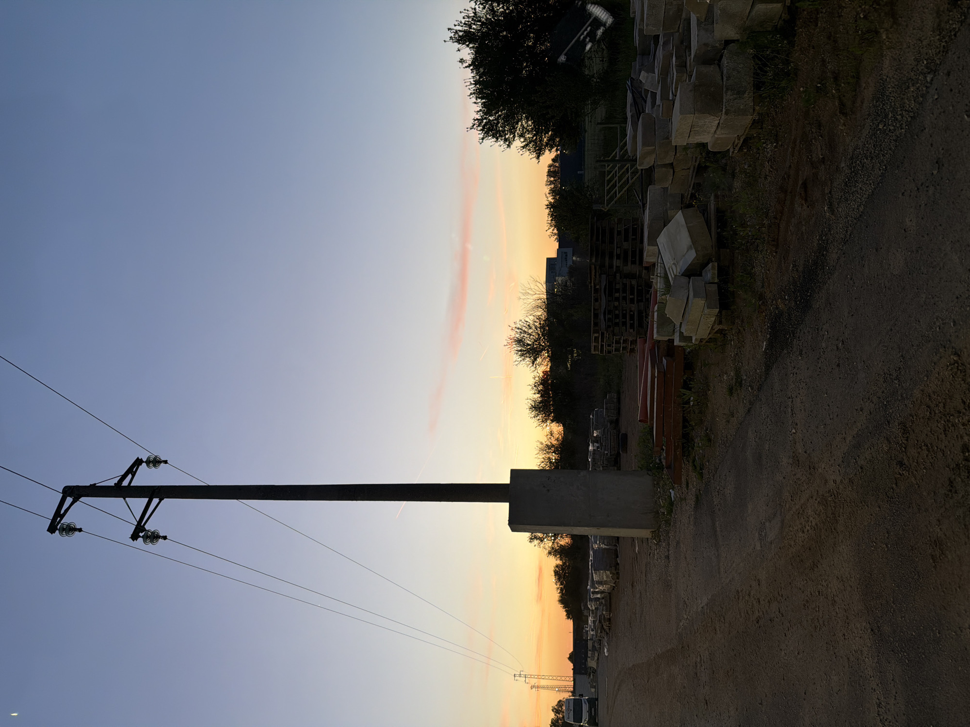 This media file presents an outdoor scene at twilight, likely sunset, in Sofia, Bulgaria. The image captures a landscape blending elements of nature and human infrastructure.
In the foreground, a tall, dark utility pole, possibly made of concrete, stands prominently on the left side, extending upwards. It features insulators and power lines stretching horizontally across the sky. The base of the pole is anchored by a solid concrete block.
To the right, a dirt or gravel path traverses the lower portion of the frame. Along this path, numerous concrete or stone blocks are stacked in an orderly fashion, suggesting a storage or construction site. Beyond these materials, a line of dark trees forms a silhouette against the horizon, with some distant buildings or industrial structures faintly visible behind them.
The sky is a striking feature, displaying a beautiful gradient. It transitions from a soft, clear blue at the top to a warm orange and yellow glow closer to the horizon, characteristic of twilight. A few thin, wispy pink clouds are present within the warmer sky tones, along with a single faint contrail.
No people are visible, and no specific activity or event appears to be taking place, creating a serene and still atmosphere. The scene evokes a sense of calm and the quiet beauty of the evening or early morning. There is no discernible text present anywhere in the image.