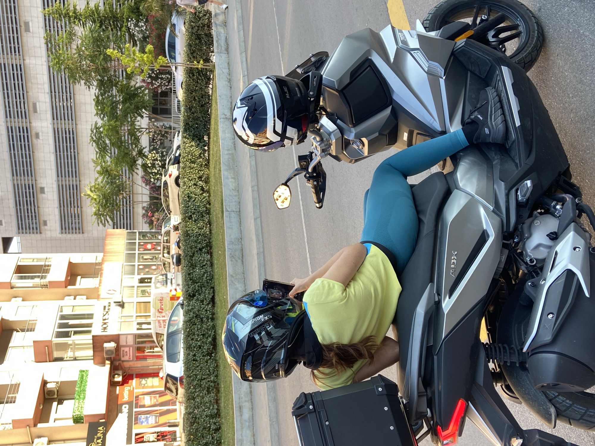 In the city of Budva, Montenegro, a person wearing a yellow t-shirt and teal leggings, along with a black and green helmet, is seated on a silver scooter. The person is holding a mobile phone and appears to be taking a picture or recording a video. Another helmet, black and white with the HJC logo, is placed on the handlebars of the scooter. In the background, there are buildings, trees, and parked cars. The scooter is parked on a street with a yellow line marking. A black storage box is attached to the rear of the scooter.