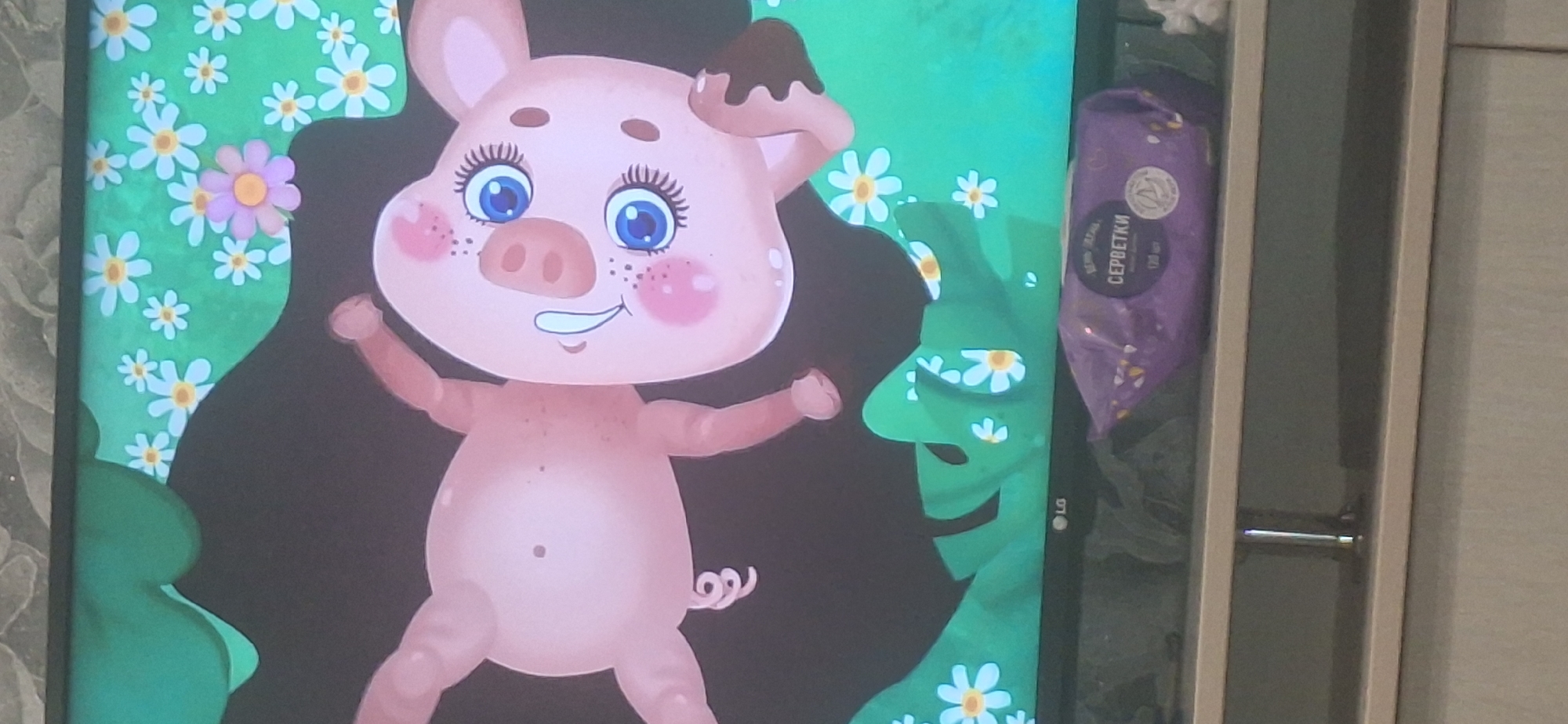 The image displays a cartoon pig lying on a dark background, with a green, floral scene framing its head and legs. The pig has large blue eyes, pink cheeks, and a curly tail. A purple package of wet wipes with Russian text, including the word "СЕРВЕТКИ" (servetki), is positioned on a shelf below the screen. The screen displays the LG logo on the bottom left. The scene appears to be indoors, with a shelf and a wall visible in the background. There is no indication of the location being in Bila Tserkva, Ukraine.