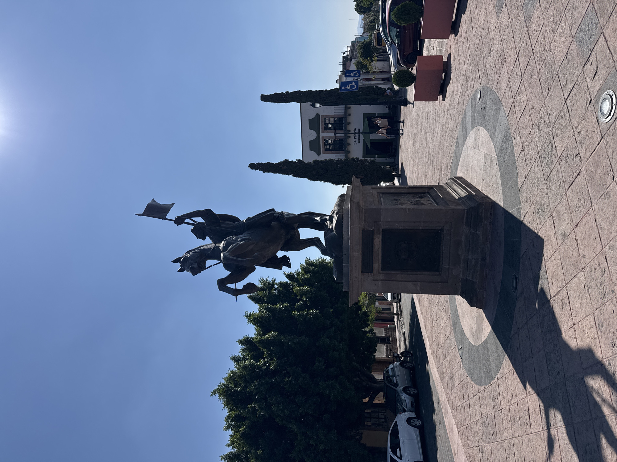 The image shows an equestrian statue in a plaza in Querétaro, Mexico. The statue depicts a man on horseback, holding a flag. The statue is mounted on a stone pedestal. In the background, there is a building with the name "La Traviata Restaurante" visible on it. Several cars are parked along the side of the plaza. The plaza is paved with light red stone tiles. The sky is a clear blue.