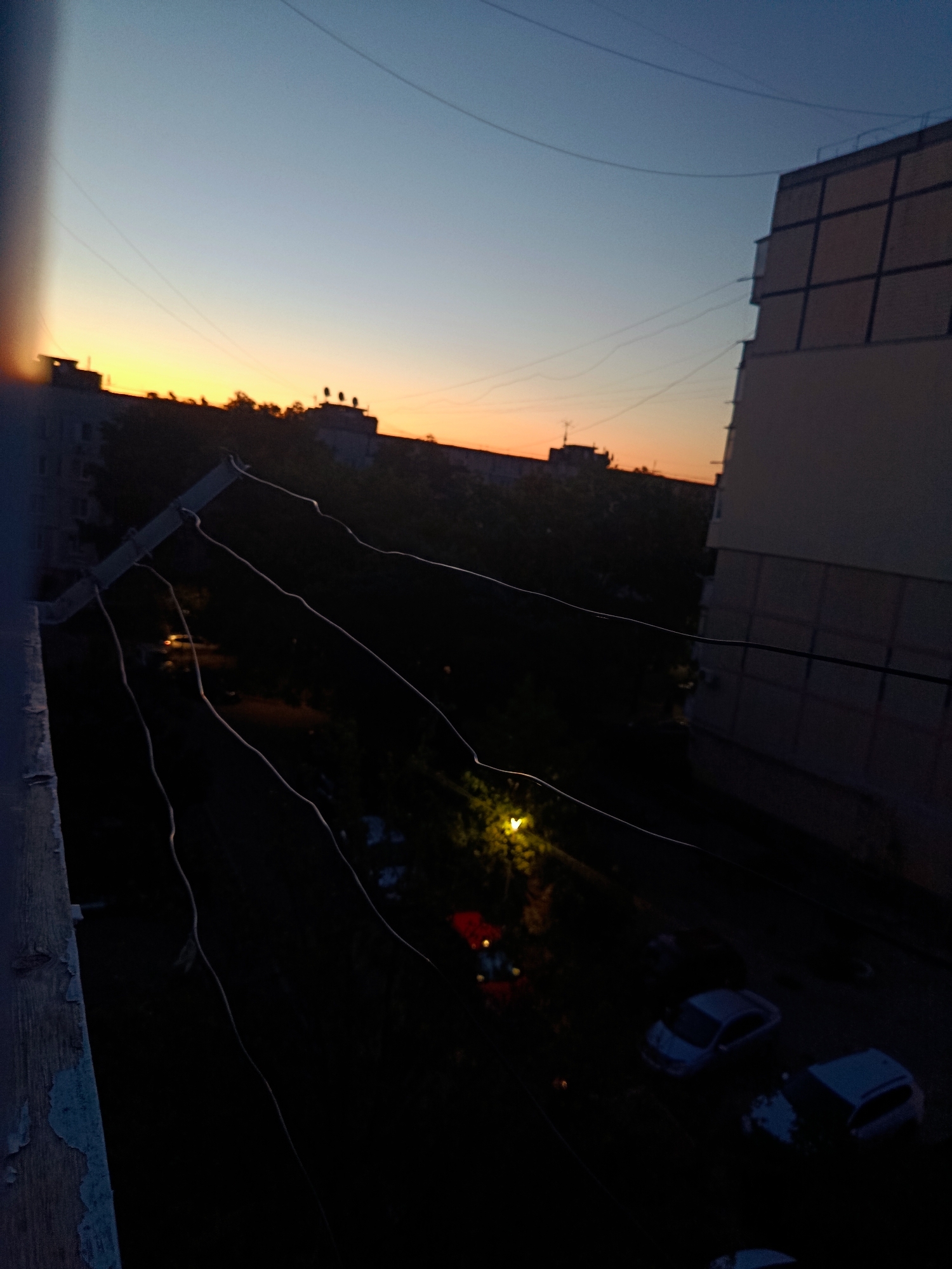 This is a photograph taken from a high vantage point, looking down onto an urban environment at dusk. The sky displays a gradient of colors, transitioning from a deep blue at the top to a bright orange and yellow near the horizon. Silhouetted against the sky are the tops of buildings and trees.
In the foreground, the edge of a structure, possibly a balcony railing, is visible on the left side. Several dark wires or cables stretch across the frame, creating a visual barrier. Below these wires, the scene is largely in shadow, with hints of green from foliage and occasional specks of light suggesting streetlights or illuminated windows.
Several vehicles are parked in the dimly lit area below, their shapes partially discernible. The overall impression is one of a quiet evening in a residential area. The image is consistent with a scene from Cherkaske, Ukraine, given the architectural style of the buildings and the urban setting. There are no people or discernible human activity in the frame.