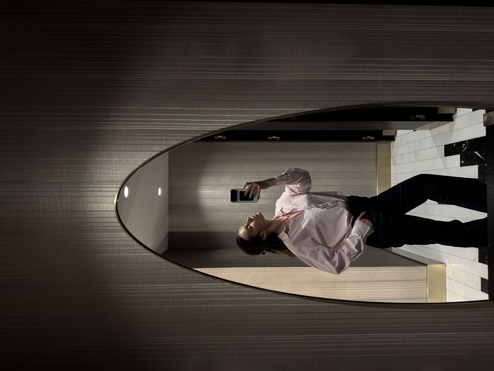 The image depicts a woman taking a mirror selfie. The woman is wearing a light pink shirt and dark pants. She is holding a smartphone in her right hand. The mirror is ovular and mounted on a wall with horizontally-lined dark wallpaper. The reflected scene shows a hallway with light-colored walls and patterned flooring. The location is unidentified in the city of Budva, Montenegro.