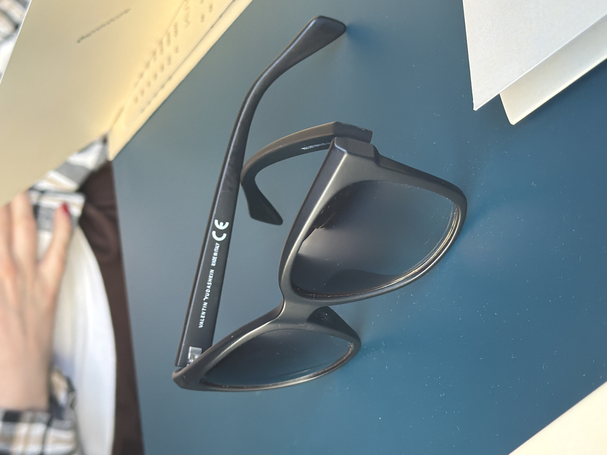 The image shows a pair of black sunglasses on a blue surface. The glasses are partially folded, with one arm extended. The brand name "VALENTIN YUDASHKIN MADE IN ITALY" is visible on the arm. A hand with red nail polish is visible near a white plate and a brown surface. The background includes a yellow book or notebook and white papers. The location in the city of Hrodna, Belarus cannot be determined from the image.
