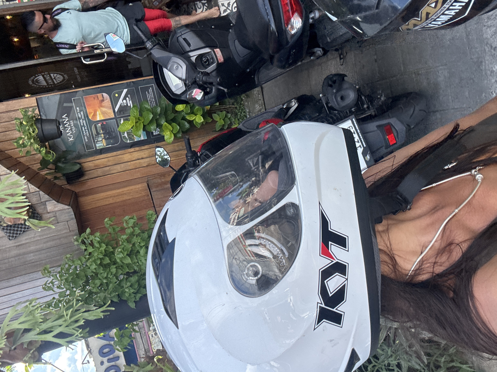 The image captures a scene in Ungasan, Indonesia, featuring a white KYT motorcycle helmet as the prominent subject. A portion of a woman with long, dark hair and a silver necklace is visible. A motorcycle is parked behind the helmet. Further in the background, there is a man, with dark hair, wearing a light-colored shirt and red shorts. He is leaning against a wooden-clad building with a sign that reads "KANANA VILLAGE" advertising gym, sauna, and pool facilities. Plants adorn the building's exterior. Another parked motorcycle is partially visible.