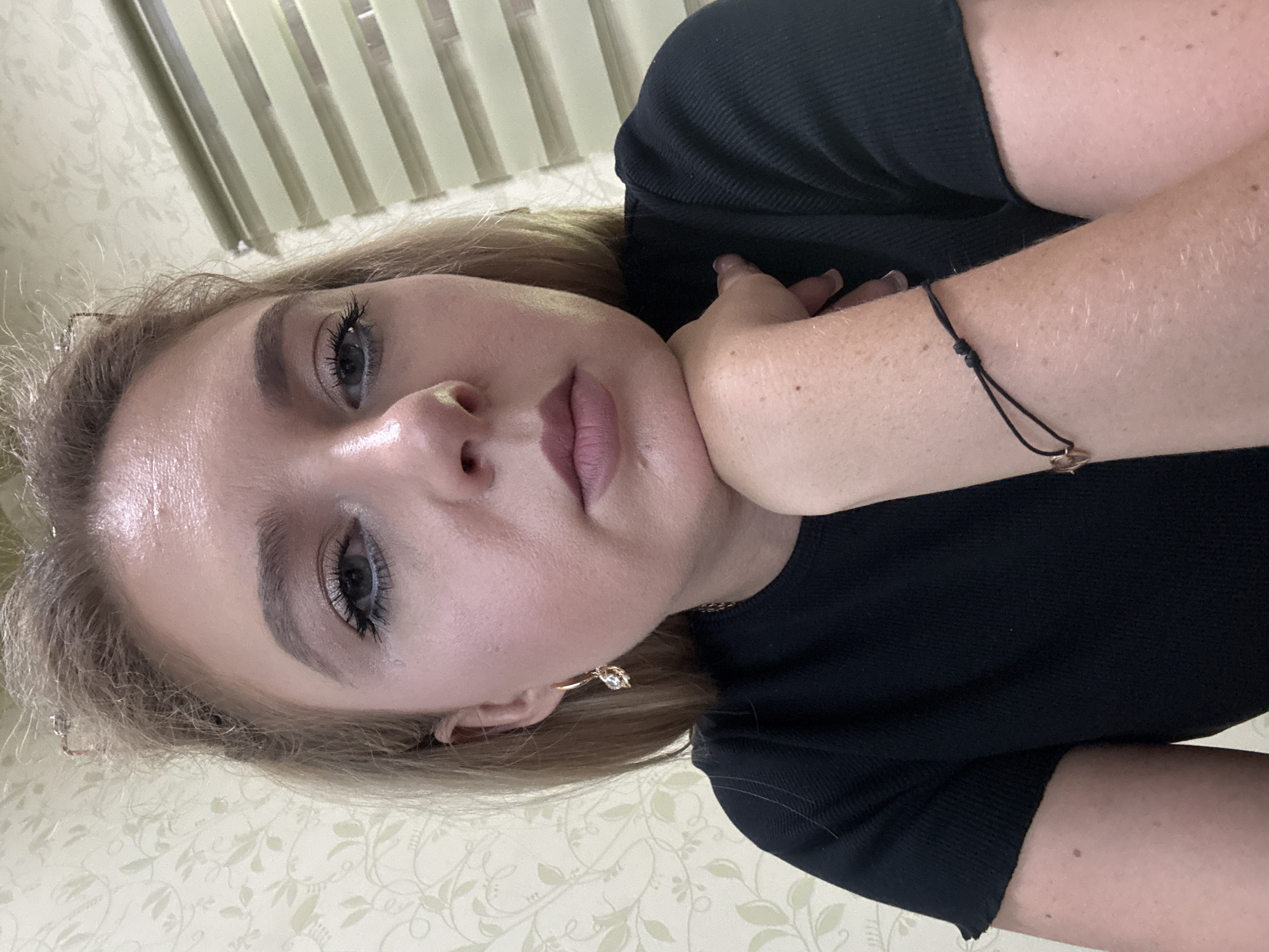 The image is a close-up selfie of a woman with blonde hair, wearing a black top and a gold earring. Her hand is resting on her chin, and she is looking directly at the camera. She is wearing makeup, including eyeshadow and lipstick. A black string bracelet with a small gold charm is visible on her wrist. The background appears to be a wall with a floral pattern and blinds. The lighting suggests an indoor setting.