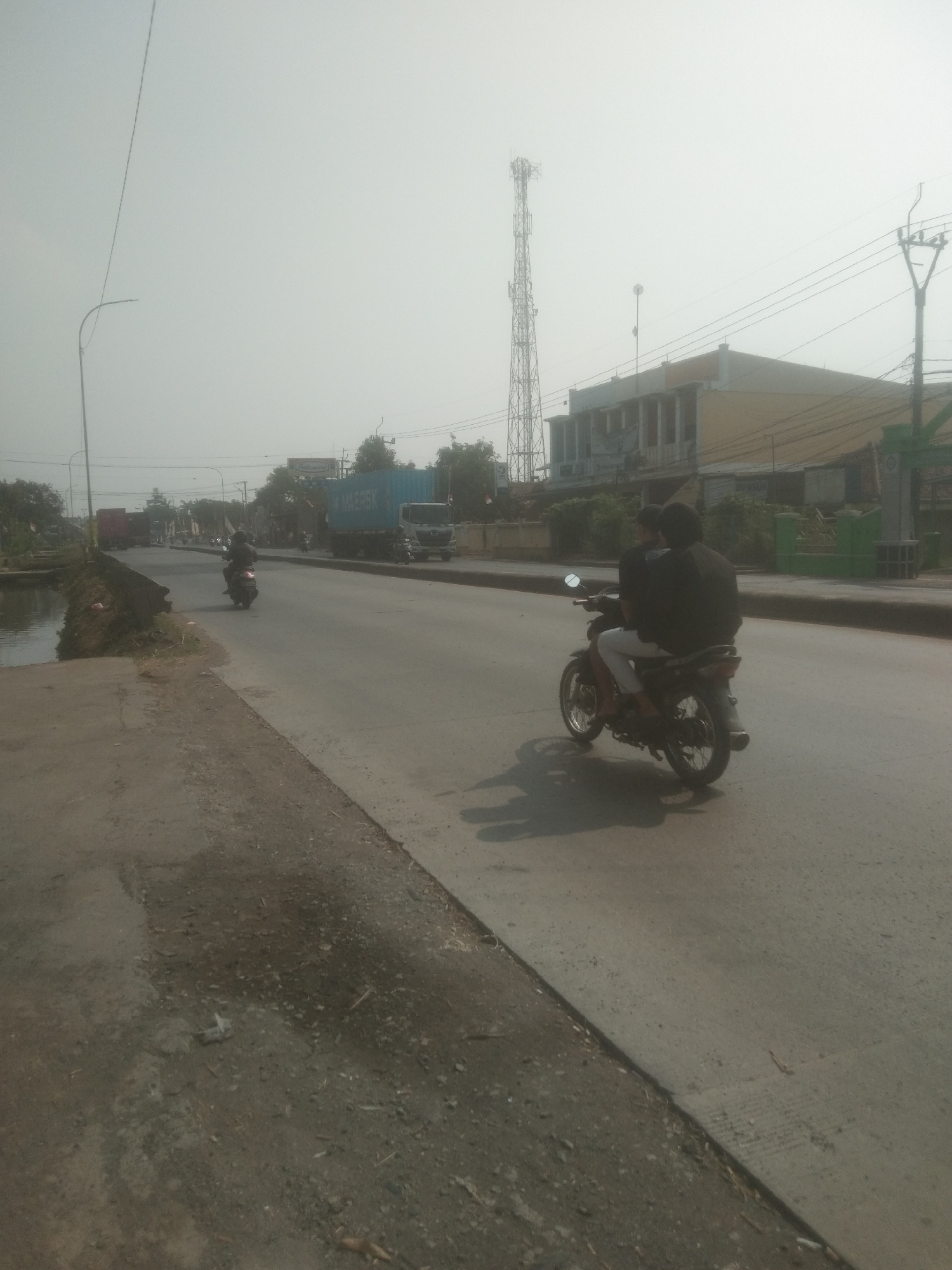 The image depicts a street scene in Subang, Indonesia, characterized by hazy atmospheric conditions. In the foreground, a paved road is visible, with a rougher, unpaved embankment to the left, bordering a small body of water. Two individuals are riding a motorcycle on the right side of the road, moving away from the viewer. The motorcycle is dark-colored, and the riders are wearing dark clothing. Further down the road, other vehicles, including trucks and motorcycles, are present. In the background, there are buildings, some with signage that appears to indicate businesses. A prominent telecommunications tower stands tall against the overcast sky. The overall environment suggests a semi-urban or industrial area on a bright, yet obscured, day.