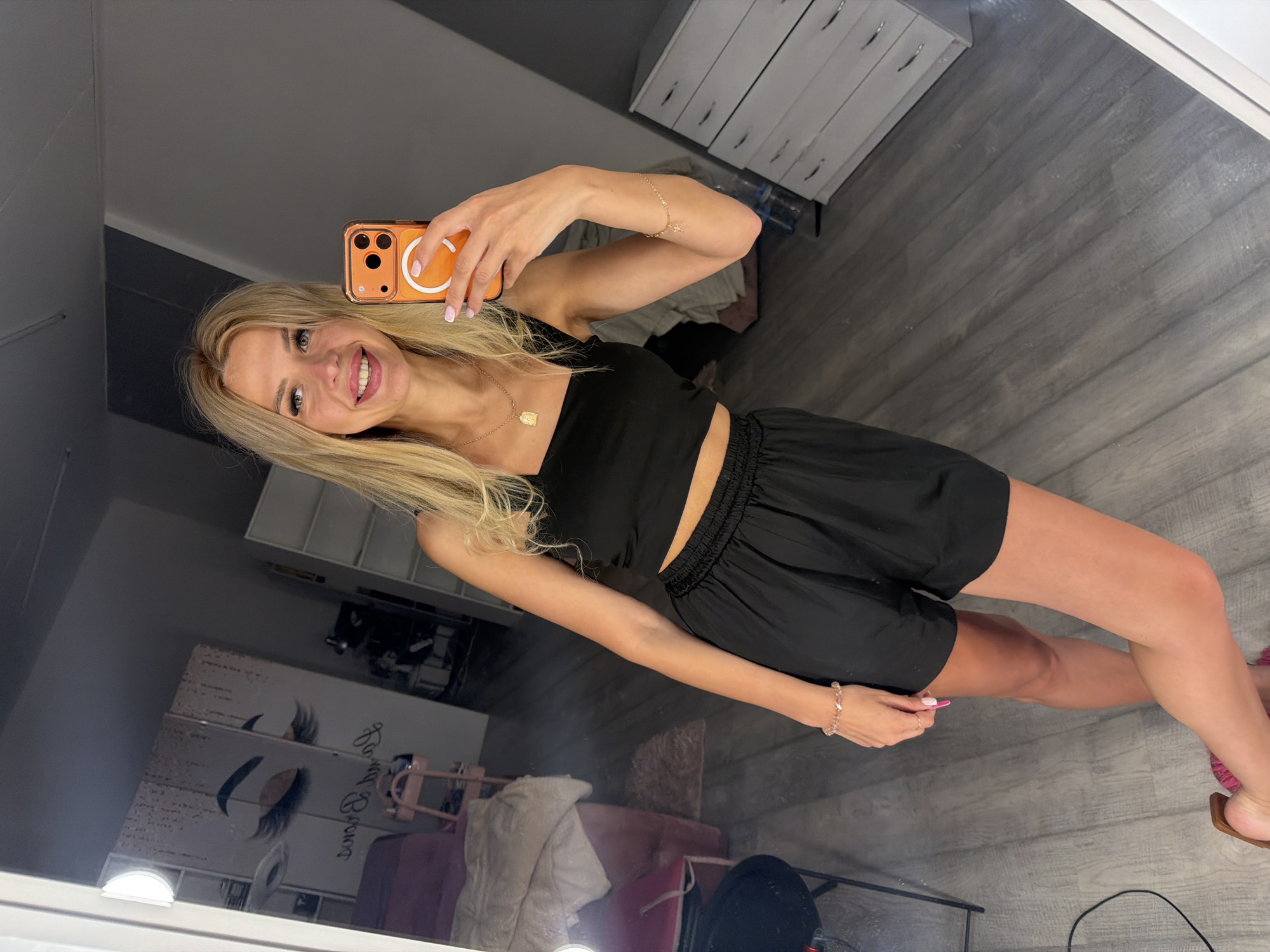A young woman with blonde hair and blue eyes, wearing a black crop top and black shorts, smiles at the camera while holding an orange smartphone. She is in a room with grey wooden floors and a white dresser in the background. A mirror reflects the woman and the room, showing a pink sofa and shelves with decorative items. The overall scene appears to be a home or salon environment in Kyiv, Ukraine.
