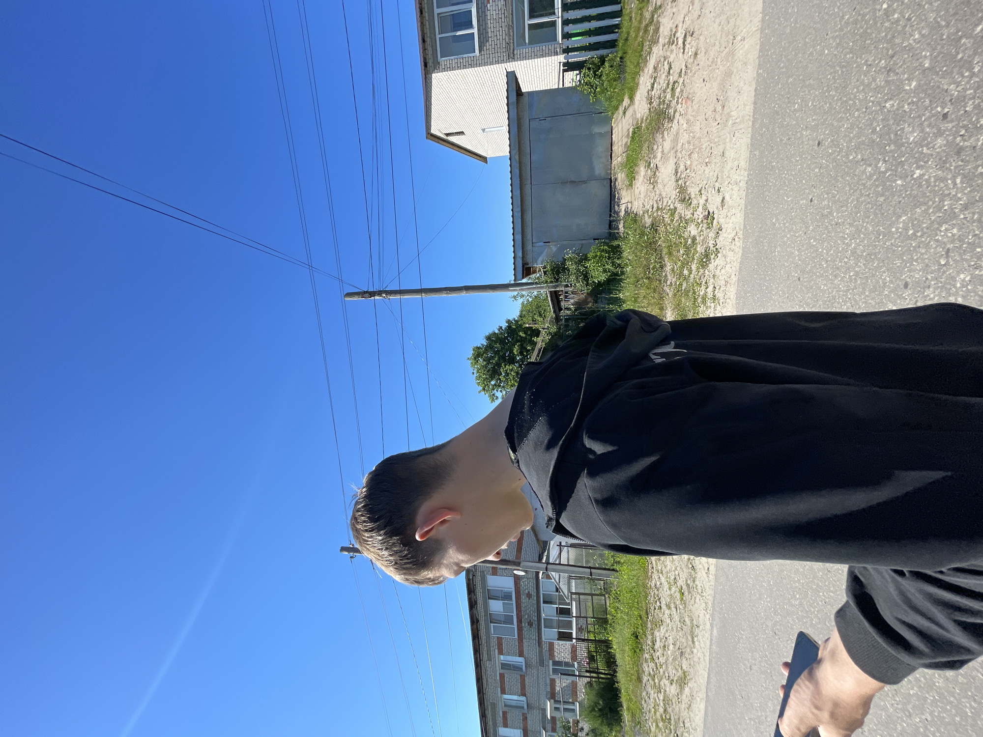 An outdoor, medium shot captures a young Caucasian male from the back and side, wearing a black hooded sweatshirt. He is looking down at a black smartphone held in his left hand. The background depicts a bright, clear blue sky with several overhead power lines. To the left is a light brick, two-story building with multiple windows. In the center and to the right are a gray metal garage with double doors and another light brick, two-story building. The foreground shows a paved road transitioning to a dirt path, with overgrown grass and foliage flanking the path. The overall setting appears to be a residential area in the city of Krasnye Baki, Russia, on a sunny day.