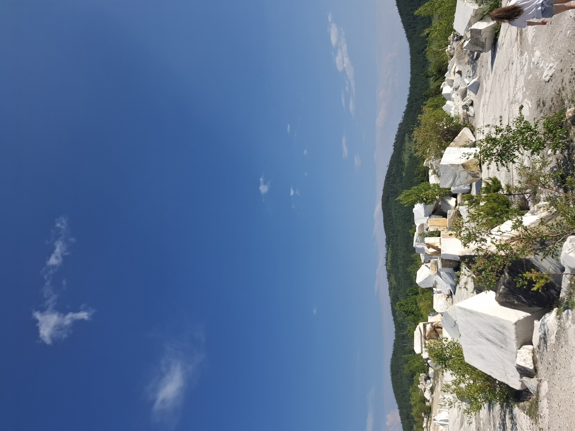 The image depicts a sunlit outdoor scene at a marble quarry in Bugul'deika, Russia. In the foreground, large, irregularly shaped blocks of white marble are scattered across a grey, rocky terrain. Green foliage, including small trees and bushes, is interspersed among the marble. In the middle ground, more marble blocks continue to be visible, with a person walking away from the camera on the right side of the frame. The person is wearing a white t-shirt and denim shorts. The background features a rolling, tree-covered hillside under a vast blue sky with scattered white clouds. The overall impression is of an industrial or natural landscape being utilized for stone extraction.