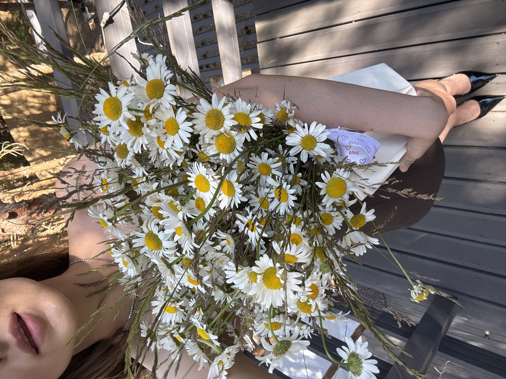 A woman is holding a large bouquet of daisies and grass. She is wearing a brown dress and black pointed-toe heels. The bouquet is wrapped in white paper with a purple ribbon that says "LOVE". She is standing on a wooden deck outdoors. The background appears to be a natural setting with trees and sunlight.