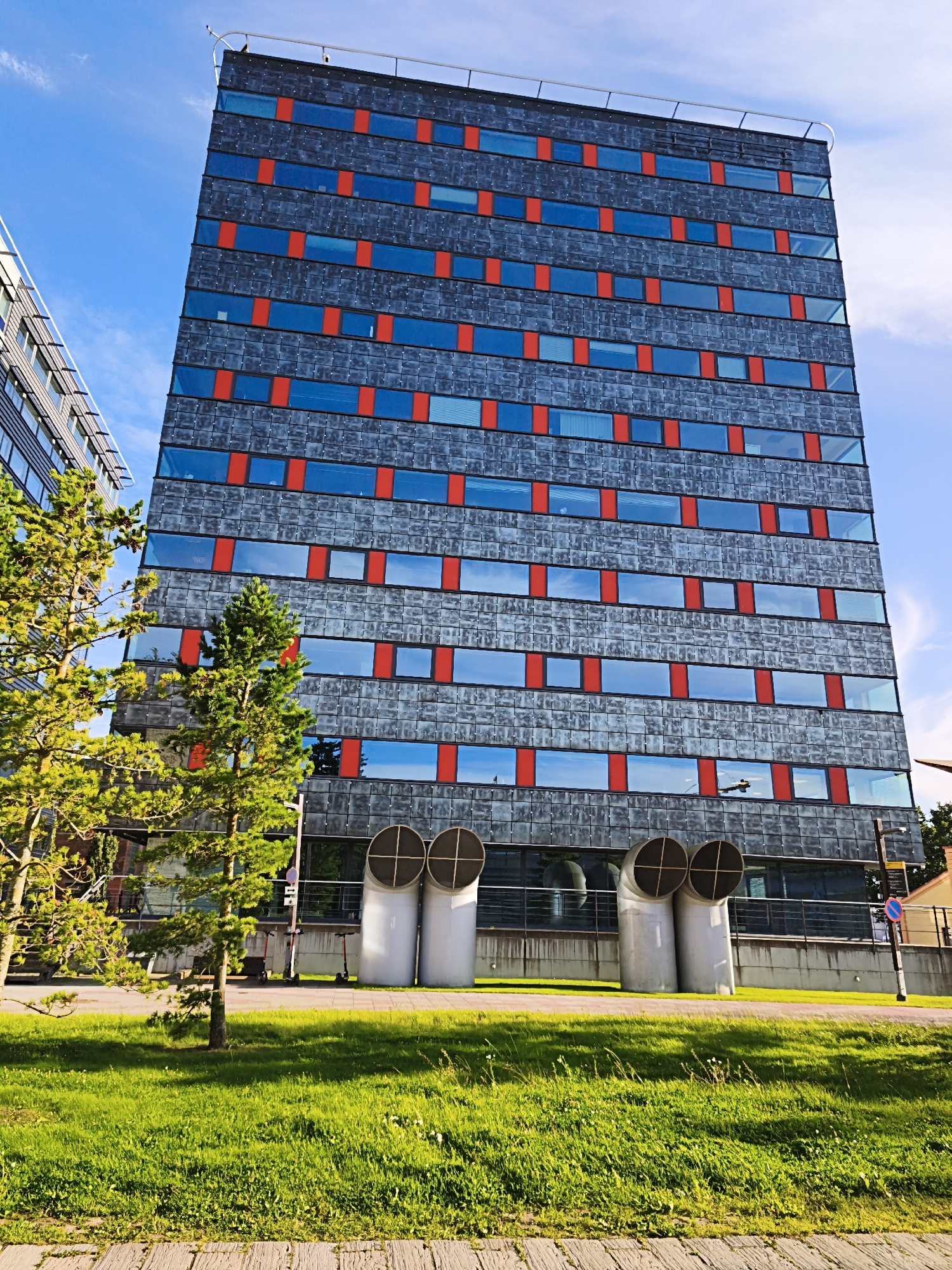 This is a wide-angle, eye-level, outdoor, daytime, vertical photograph of a modern, multi-story building in Tallinn, Estonia. The building's facade is predominantly gray with horizontal bands of blue windows and interspersed vertical red accents. Two large, white, cylindrical ventilation pipes are visible at the base of the building. In the foreground, a vibrant green lawn with scattered white flowers is illuminated by sunlight. Two young pine trees stand in front of the building, to the left of the frame, their branches casting shadows on the grass. In the background, a portion of another building with a glass facade can be seen to the left, and trees and a small building with a brown roof are visible to the right. Two electric scooters are parked near the ventilation pipes. The sky is bright blue with scattered white clouds.