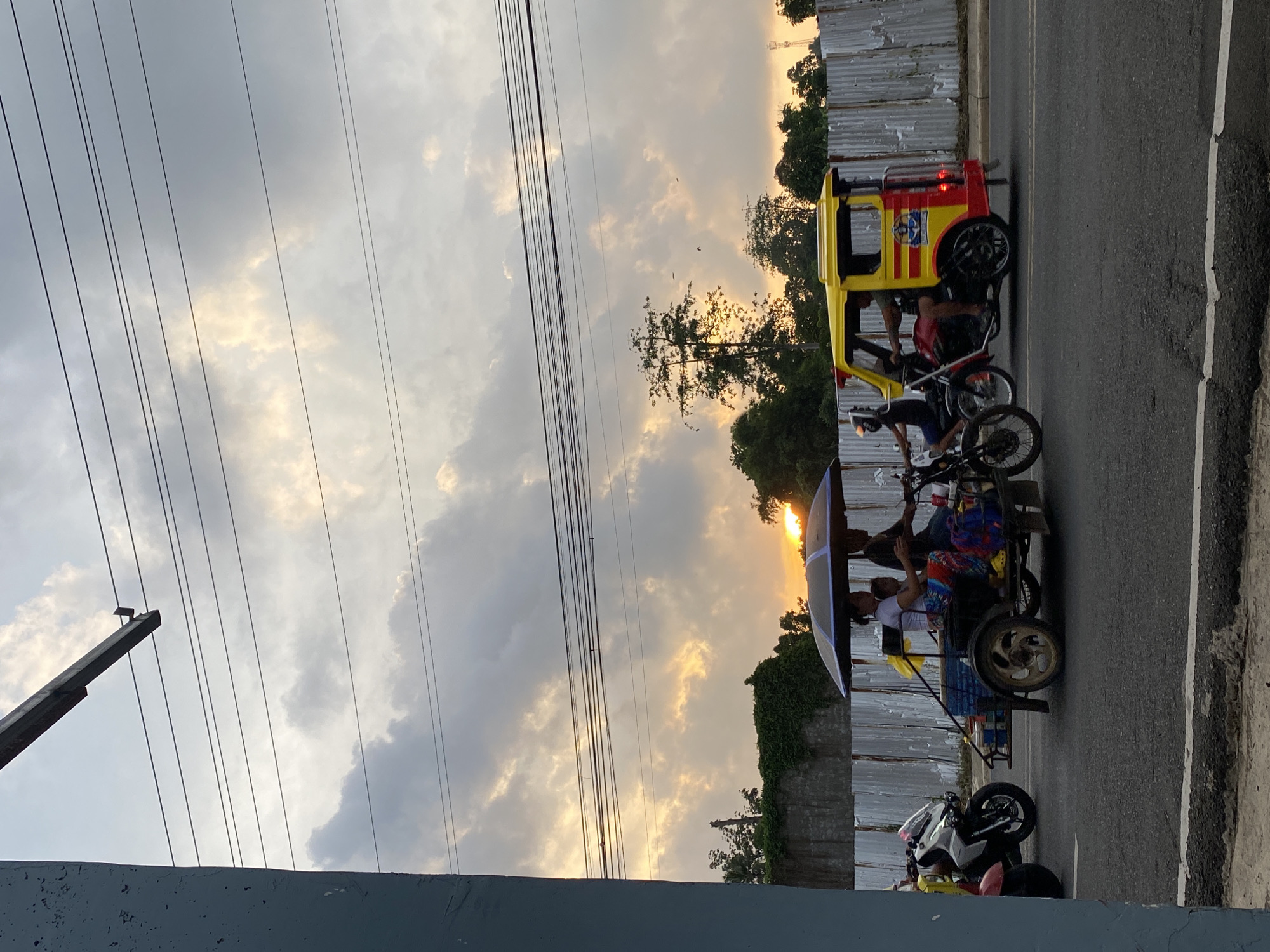 The image captures a street scene at sunset in Davao City, Philippines. The sky is largely overcast with clouds, some illuminated from beneath by the setting sun, casting warm golden light through breaks in the cloud cover. Multiple dark power lines extend horizontally across the upper frame, supported by a metal beam partially visible from the top left. A large, dull blue-gray concrete pillar or wall occupies the far left of the frame, from top to bottom.
On an asphalt road, several vehicles are in motion, heading from left to right. In the mid-ground, a dark-colored motorcycle with an attached sidecar, functioning as an open tricycle, carries multiple passengers and cargo. An open dark umbrella provides shade over the sidecar, where an adult female in a white shirt and patterned skirt, an adult male driving the motorcycle, and at least one child are visible. Blue containers are also present in the sidecar. Partially obscured behind this tricycle is a white and pink scooter or motorcycle. Further to the right, a yellow and red tricycle, with an enclosed sidecar featuring a decorative emblem, is also moving. A driver and at least one passenger are visible inside this vehicle.
In the background, a weathered corrugated metal fence stretches horizontally, separating the road from an area with dense green foliage, trees, and parts of a concrete wall. More trees are visible against the horizon, with the faint silhouette of a distant structure, possibly a communication tower, on the far right. The overall scene depicts a typical evening commute or activity in an urbanized area of the Philippines.