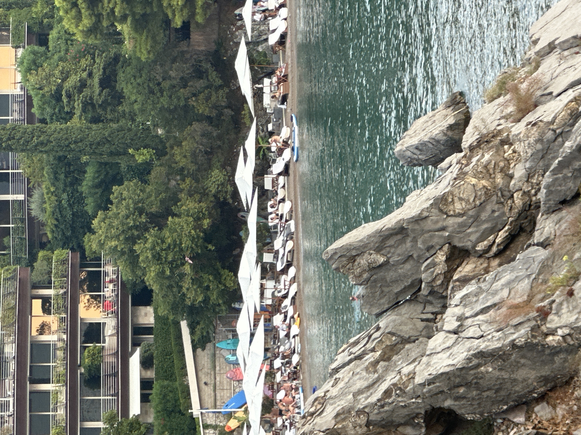 The image shows a beach in Budva, Montenegro, with a resort building in the background. The beach is lined with lounge chairs and umbrellas, and several people are visible relaxing on the sand and in the water. Surfboards and paddleboards are propped up against a wall on the left side of the image. The water is a deep blue-green, and the rocky shoreline is in the foreground.