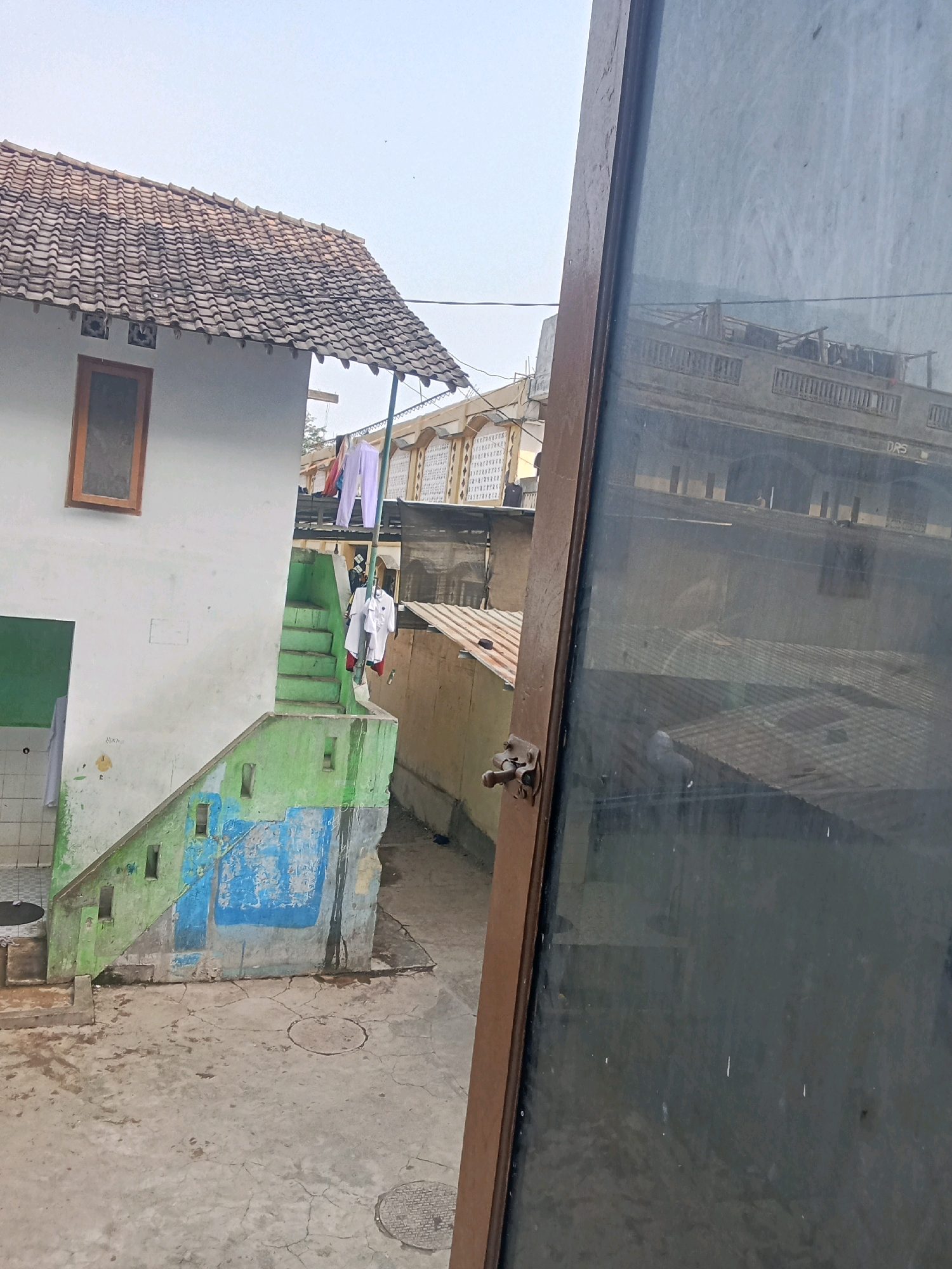 In the city of Sumberadi, Indonesia, an outdoor scene features a white building with a tiled roof, featuring a wooden-framed window. Adjacent to it, green-painted stairs lead up to an entrance. Several pieces of clothing, including white garments, are hung on a clothesline. In the foreground, a large, dark pane of glass, possibly from a window or door, is visible on the right, showing reflections of buildings. The ground is paved with concrete, showing cracks and some circular manhole covers. The overall environment appears to be a residential or communal area with multiple buildings and a clear sky overhead.