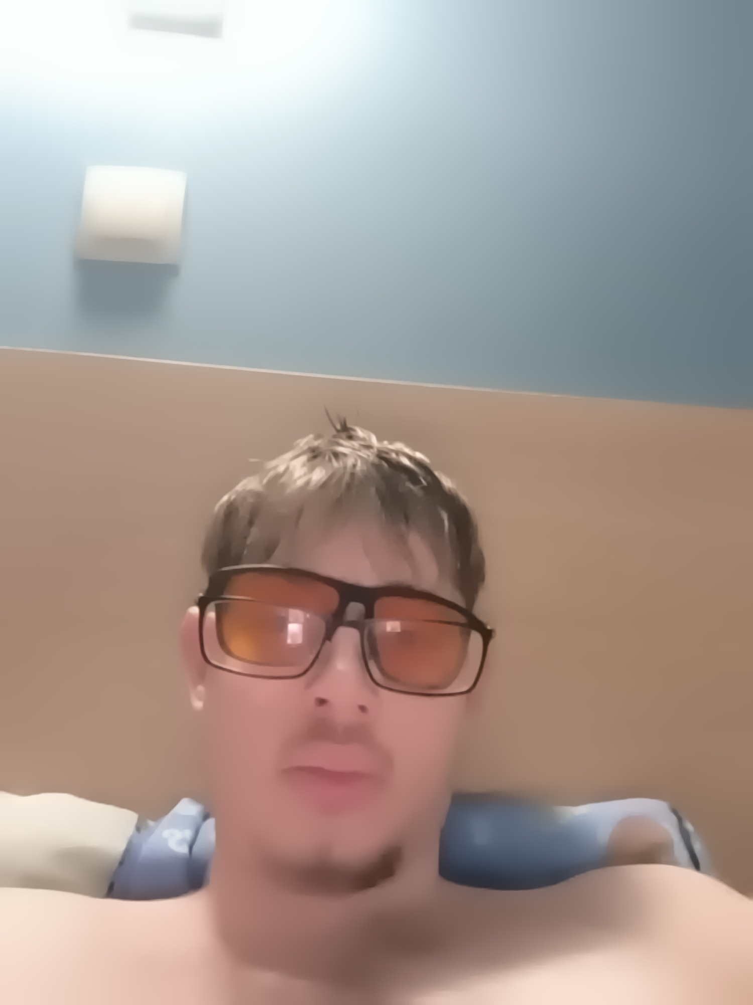A young man is depicted in a close-up, chest-up indoor shot, likely taken in Sofia, Bulgaria. He looks directly at the camera with a neutral expression. He has light-colored, somewhat messy hair, a faint mustache, and a light goatee. He is wearing prominent dark-framed glasses with distinctive orange-tinted lenses, which obscure his eyes. The man appears to be shirtless.
Behind him, a light blue pillow with a subtle white pattern is visible. The background features a two-toned wall: the upper section is light blue, while the lower section, acting as a headboard or paneling behind the man, is light brown. Two artificial light fixtures are mounted on the upper blue wall; one is rectangular and positioned higher, and the other is a square shape located below it. Both fixtures are illuminated, casting a bright, diffused light and suggesting the photo was taken indoors, possibly at night or in the evening. The overall scene conveys a casual and relaxed atmosphere. There is no visible text in the image.