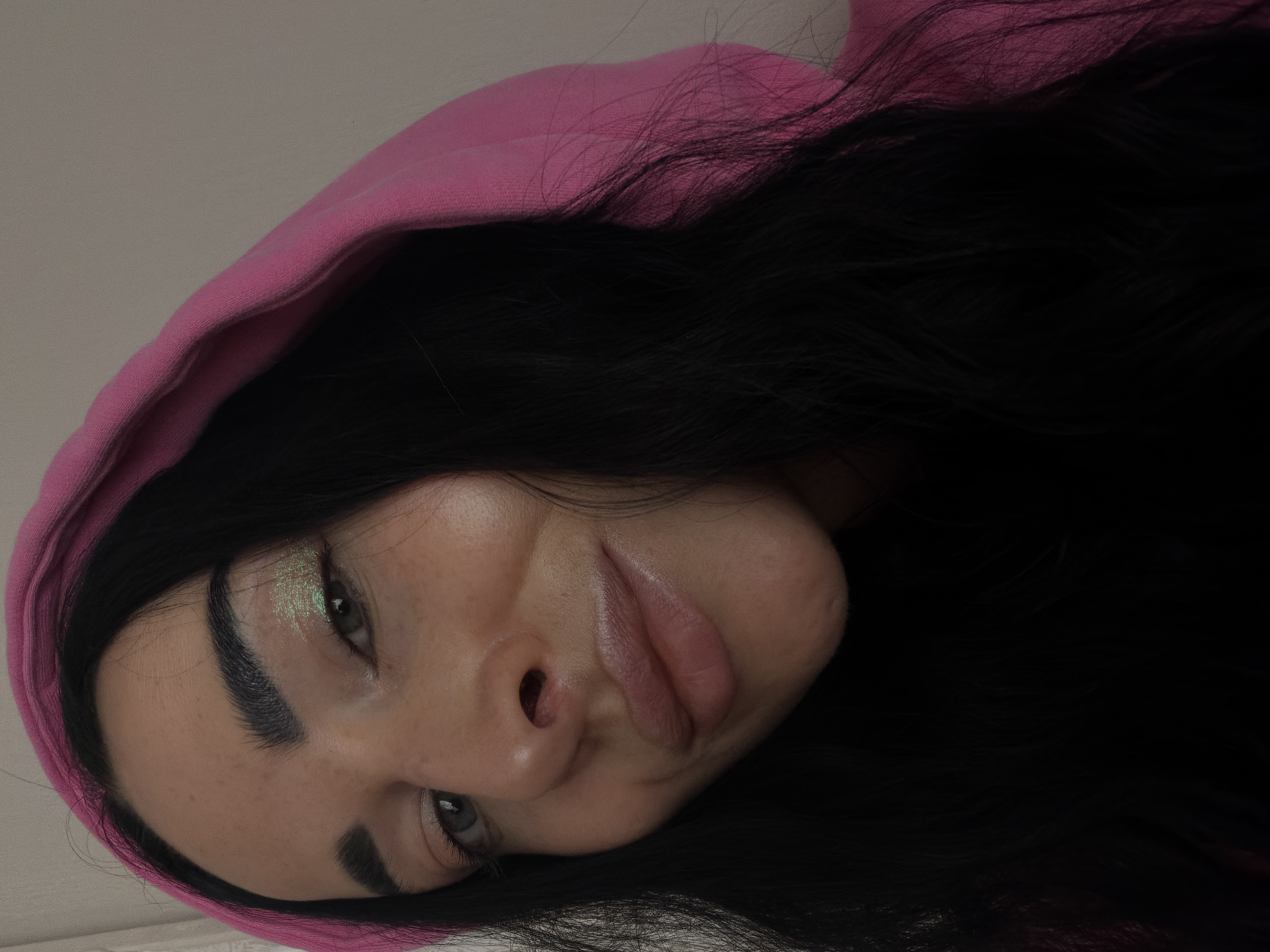 This close-up portrait, captured in Saint Petersburg, Russia, features a person, likely female, looking towards the viewer. The image is tilted, presenting the subject's face at an angle. She is wearing a vibrant pink hooded garment, with dark, long, wavy hair spilling from beneath the hood and around her shoulders. Her dark eyebrows are well-defined, and her eyes are open, looking directly at the camera. Notable makeup includes a shimmery, light green eyeshadow applied to her eyelids. Freckles are visible across her nose and cheeks. Her lips are full and slightly pursed or pouted, suggesting a casual or playful expression. The background is a plain, light color, indicating an indoor setting. No text is visible in the image.