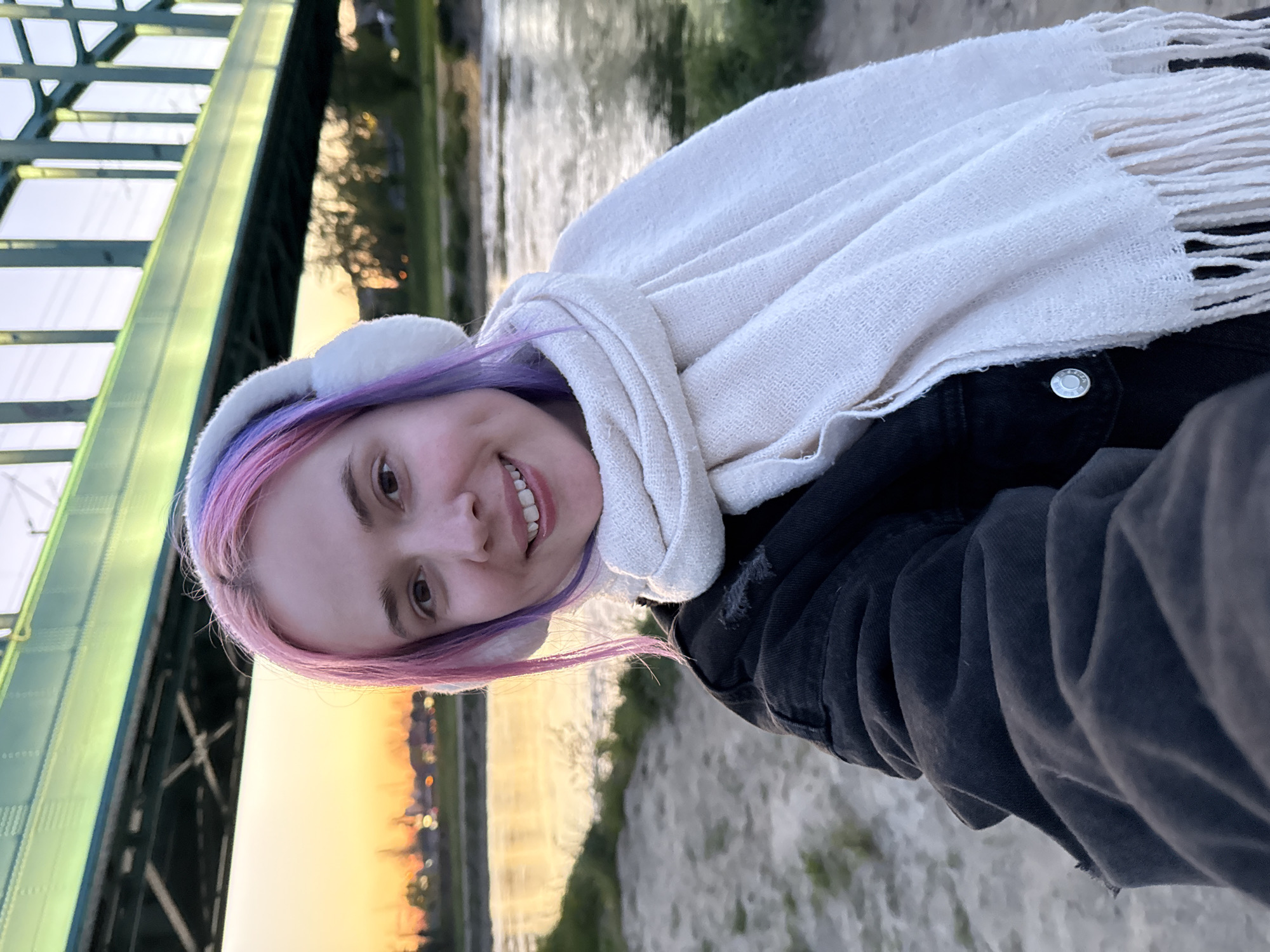 A vertically oriented medium shot depicts a woman with pastel-colored hair standing on the bank of the Sava River in Zagreb, Croatia. She is wearing white earmuffs and a white scarf. She is smiling. The background features a sunset reflecting on the river, and the green metal structure of a bridge is visible in the upper-left corner. Sandy ground and green grass are visible along the riverbank. The woman is wearing a dark-colored jacket or coat.