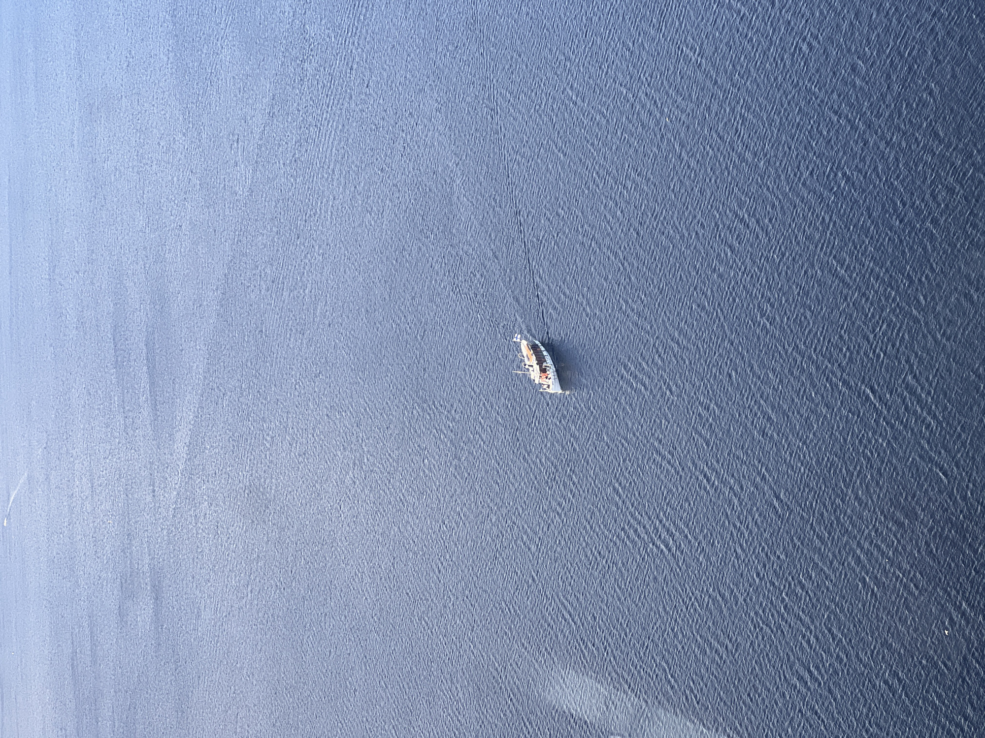 An aerial, wide shot captures a small white boat with an orange trim sailing on a calm, dark blue body of water. The boat, positioned slightly right of the center of the frame, is leaving a subtle wake behind it. The water's surface is gently rippled, creating a textured pattern across the vast expanse. The overall scene is serene and devoid of any prominent landmarks, making it impossible to determine a specific location within Tampere, Finland, or any other city.