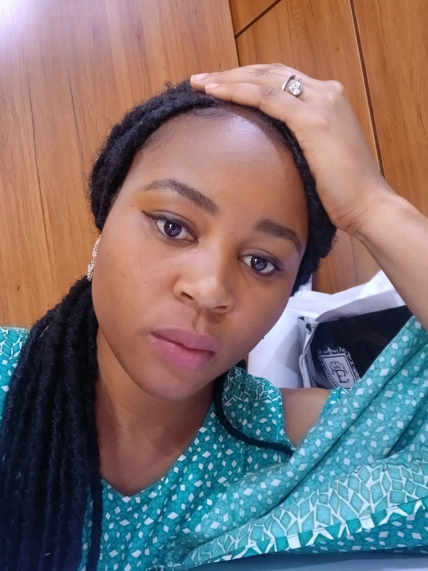 A woman is shown from the chest up, taking a selfie. She is wearing a teal and white patterned blouse and has black braids. Her left hand is raised to her forehead, and she is wearing a ring on her left index finger. Her right ear is adorned with a diamond earring. She is wearing makeup, including eyeliner and lipstick. The background consists of a wooden wall and a white bag. The lighting suggests an indoor environment. The image does not contain information indicating it was taken in Gura Top, Nigeria.