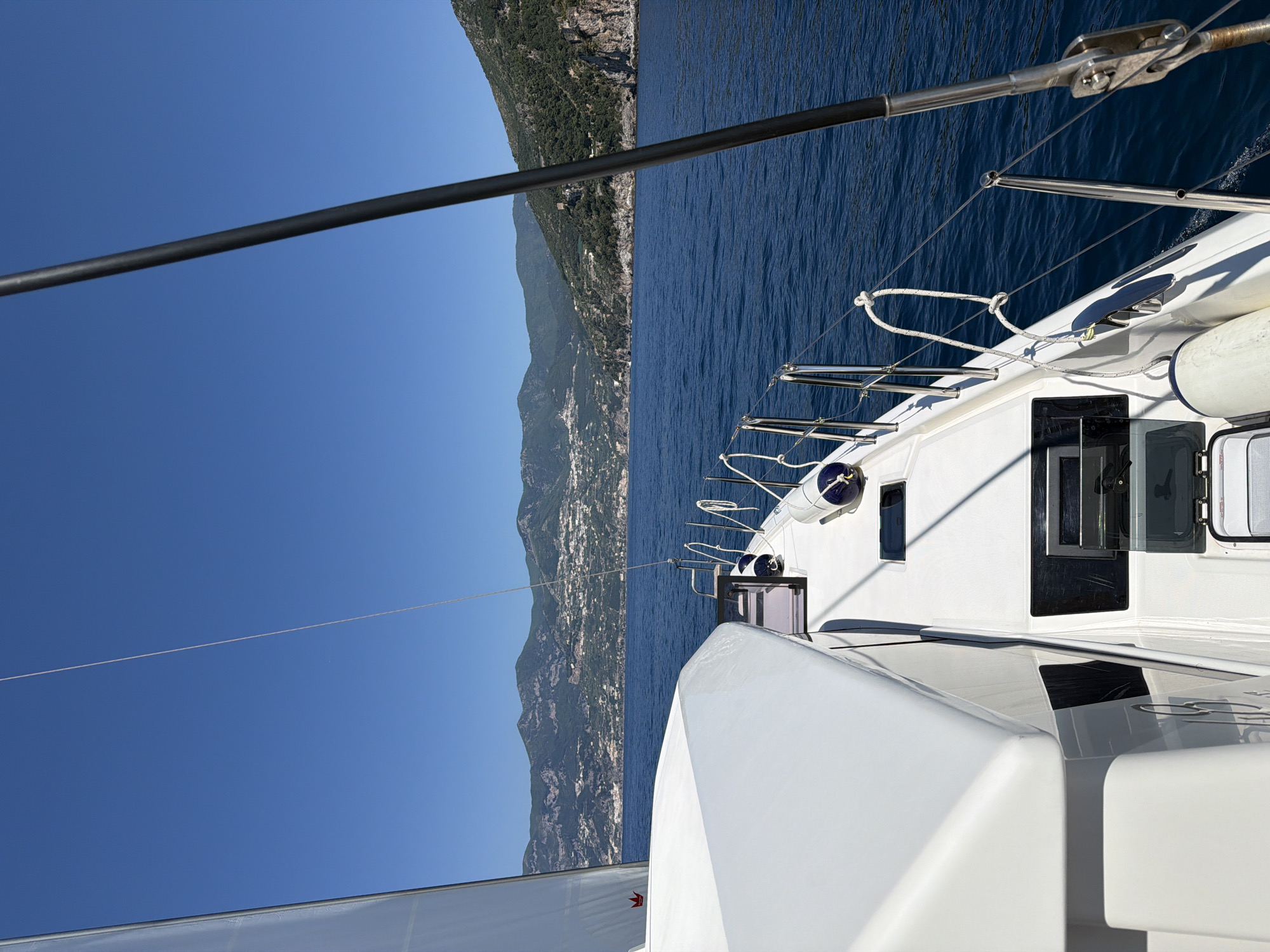 View from a boat sailing near Maiori, Italy. The boat is white with stainless steel rigging and a partial view of the sail is visible. Across the water is the coastline of Maiori, with buildings nestled on the hillside. The sky is clear blue, and the water is a deep blue.