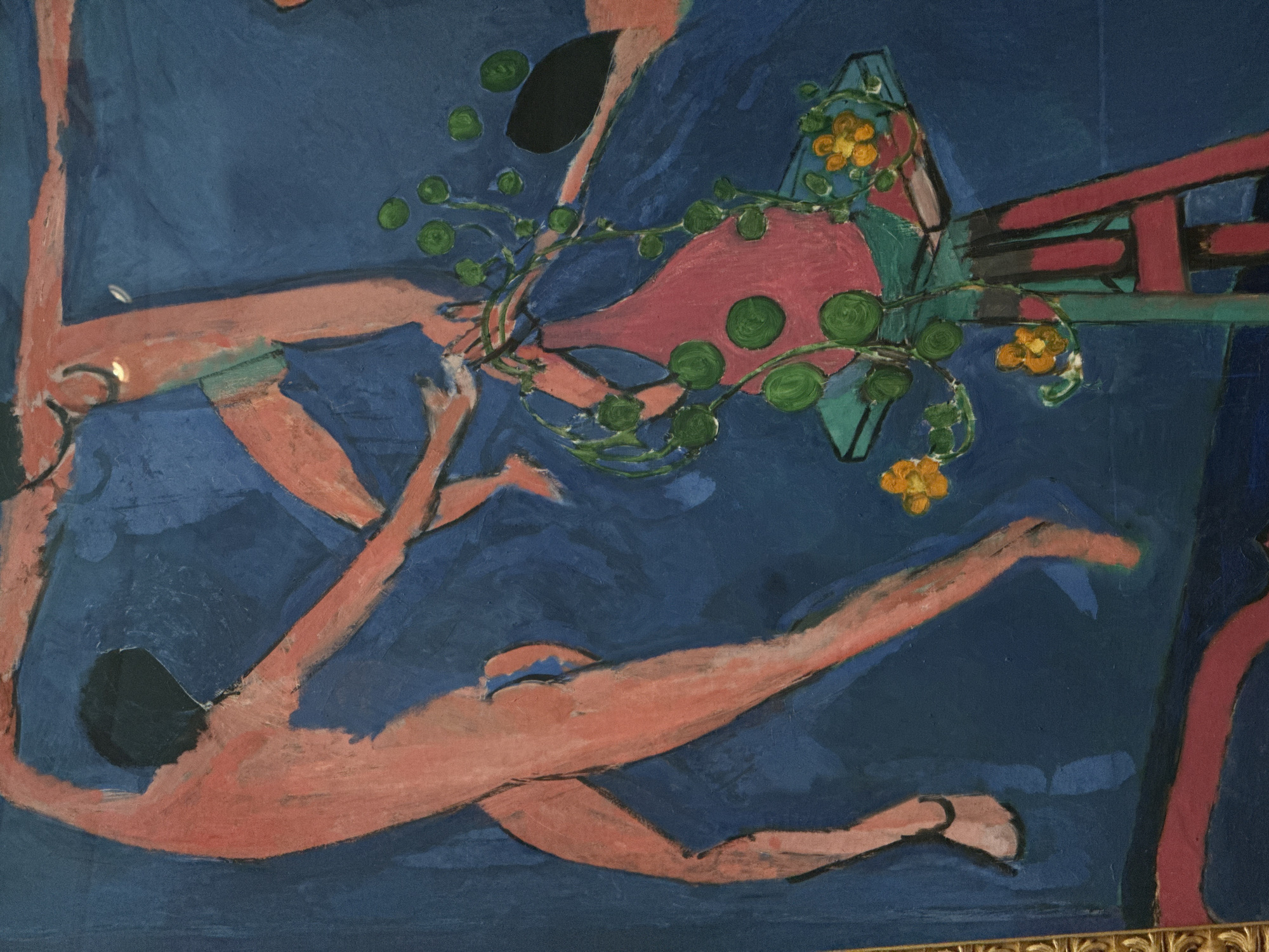 The image shows a painting with a predominantly blue background. Two nude figures, painted in shades of peach, are depicted. One figure is lying down, with arms and legs outstretched. The other figure is positioned diagonally above, appearing to hold a pink vase with green circular designs and yellow flowers. Above the vase, there is an abstract, geometric structure with red and black elements. The scene appears to be surreal and dreamlike, with an emphasis on color and form rather than realistic representation. The location in Moscow, Russia, is not discernible from the painting itself.