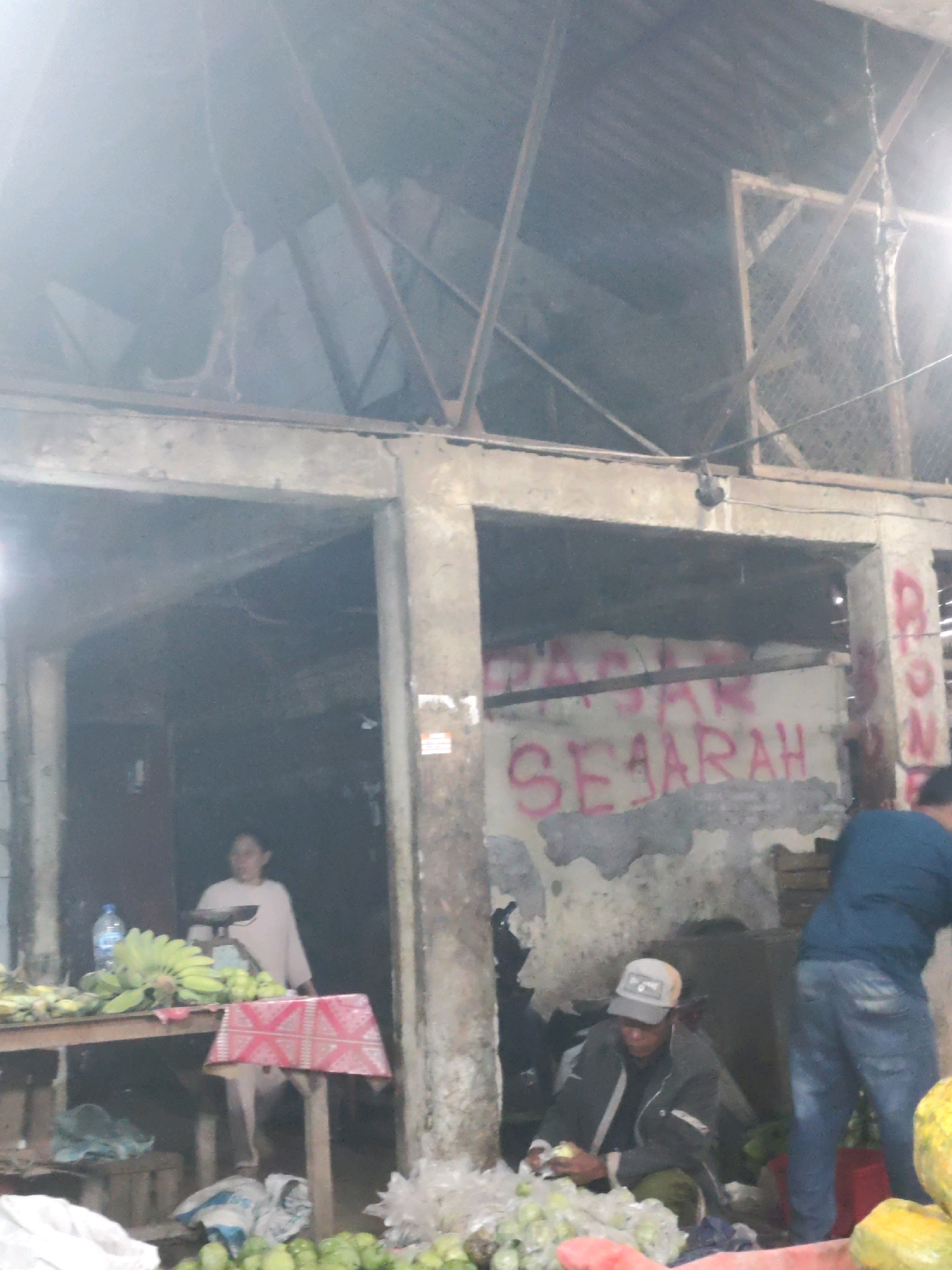 In Karawang, Indonesia, this image captures an outdoor market scene under a dilapidated roof structure. Two individuals are actively engaged with produce. In the foreground, a person wearing a cap and dark jacket is seated on the ground, sorting through a pile of green fruits and what appear to be plastic bags. To the right, another person in a blue shirt and jeans is standing and interacting with a red container. In the background, a woman in a light-colored top stands behind a table laden with bananas and other fruits, with a weighing scale visible. Graffiti, including the words "PASAR SEJARAH," is spray-painted on a concrete wall in the middle ground. The environment is characterized by concrete pillars and a corrugated metal roof, suggesting an informal or open-air market setting.
