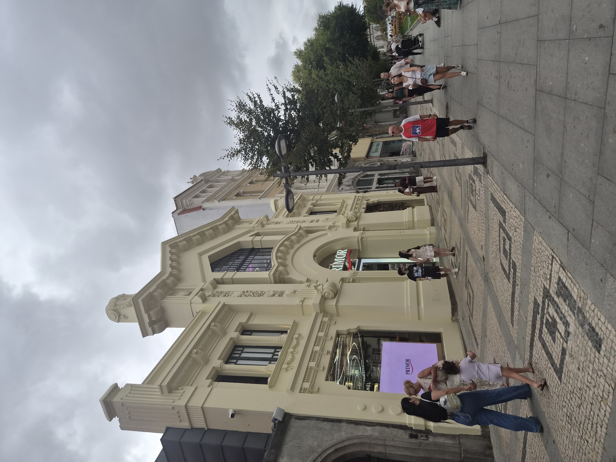 The image captures a street scene in Braga, Portugal, under an overcast sky. The foreground features a pedestrian walkway paved with intricate geometric patterns of light and dark stones. Several individuals are walking along the sidewalk. On the left side of the image, a light-yellow building with classical architectural elements dominates the scene. This building features large windows, decorative moldings, and an arched entrance. A sign for "PRIMOR" is prominently displayed above an entrance, suggesting a retail establishment. To the right of this building, other structures with varied architectural styles line the street, including a multi-story building with red-tiled roofs. Trees with lush green foliage are interspersed along the sidewalk, providing shade. In the middle ground, people are seen walking, some singly, some in pairs, and a family with a child. Further down the street, more pedestrians and a stroller are visible, indicating a busy urban environment. The overall atmosphere is that of a pleasant, though overcast, day in a European city.