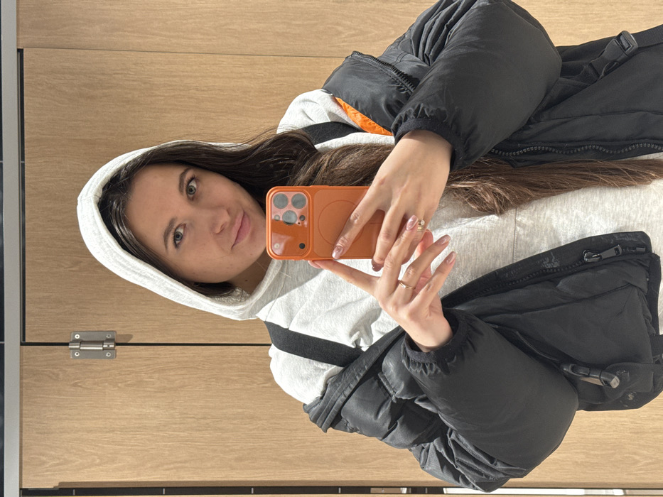 A woman wearing a light gray hooded sweatshirt and a black puffer jacket is taking a selfie in front of a wood-paneled wall with a metal door latch on the left. She's holding an orange phone in her hands, which are adorned with rings and manicured nails. Her long brown hair falls around her shoulders, partially obscuring the sweatshirt.