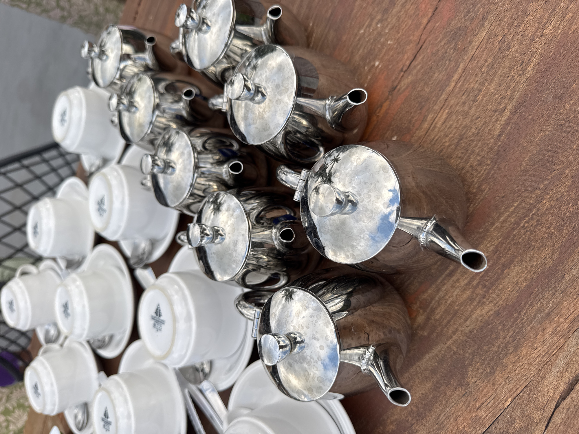 The image shows a collection of silver teapots and white teacups with saucers arranged on a wooden surface. The scene features multiple teapots with round lids, and the cups are stacked alongside them. The location is not verifiable as Rendang, Indonesia based on the visual information alone.