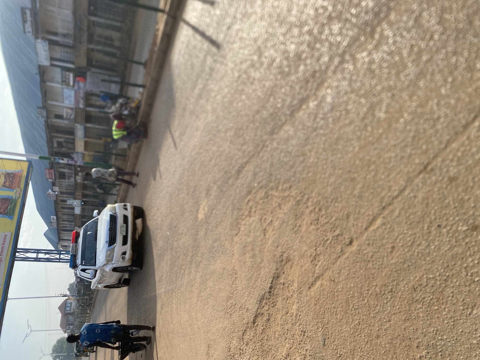 In Bukuru, Nigeria, a white security vehicle with flashing blue and red lights is parked on the right side of a dirt road. A person in a blue patterned shirt is walking away from the vehicle, pulling a wheelbarrow. In the background, several people are working, possibly cleaning the area, with one person wearing a high-visibility yellow vest. The street is lined with buildings, and there is a large billboard overhead. The sun is casting shadows, indicating it is likely daytime.
