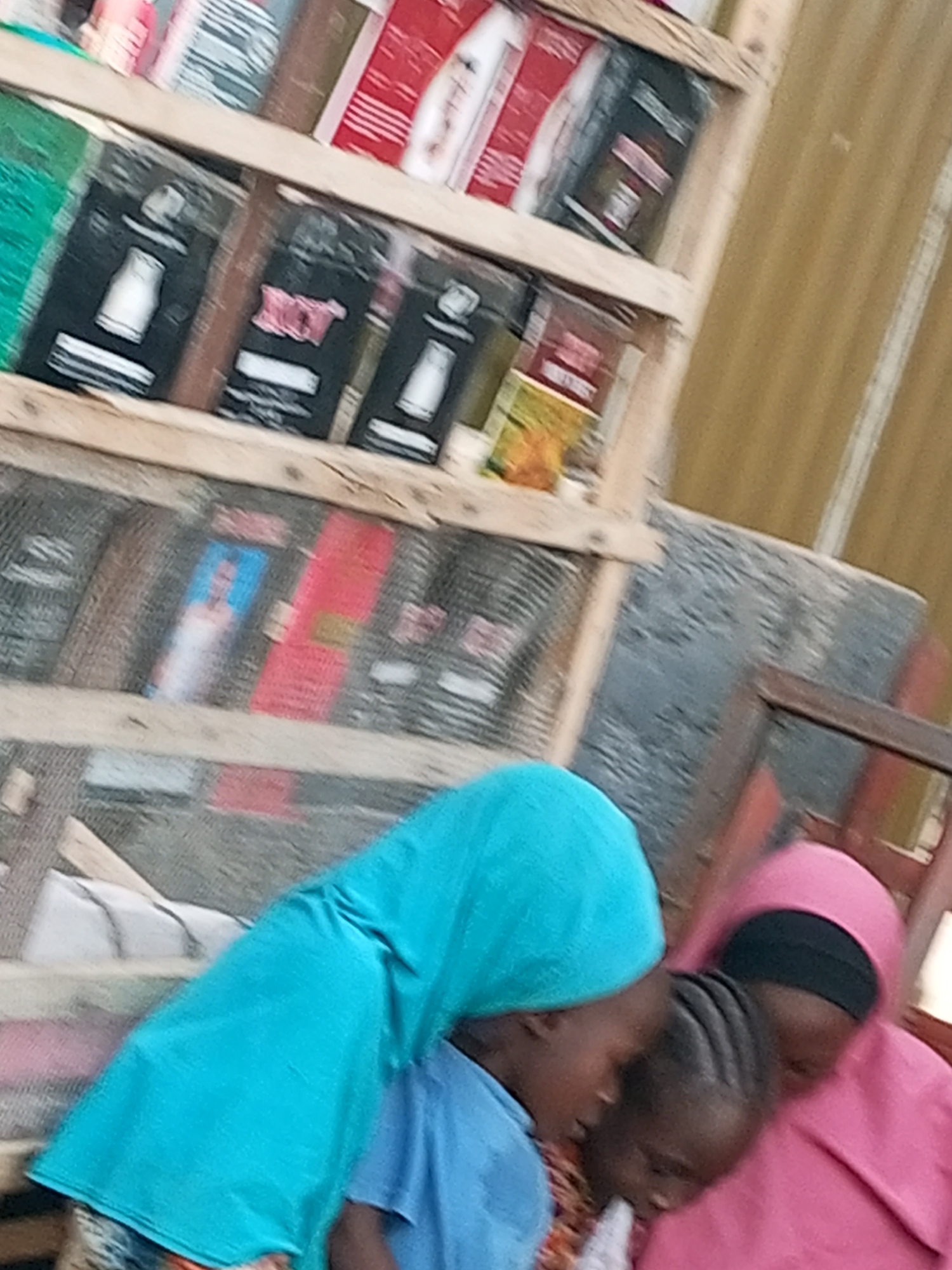 In Tempure, Nigeria, a wooden shelf displays various items, likely packaged goods, on multiple levels. The lower shelf is covered with netting and holds more items, some with visible colorful labels. In the foreground, three young individuals, two appearing to be girls wearing hijabs in blue and pink, are huddled together. The girl in the blue hijab leans towards the center, while the one in the pink hijab looks down. The third individual, positioned between them, has dark hair and is also looking downwards. They are close to each other, suggesting an interaction. The background appears to be an outdoor setting with a corrugated metal wall on the right.