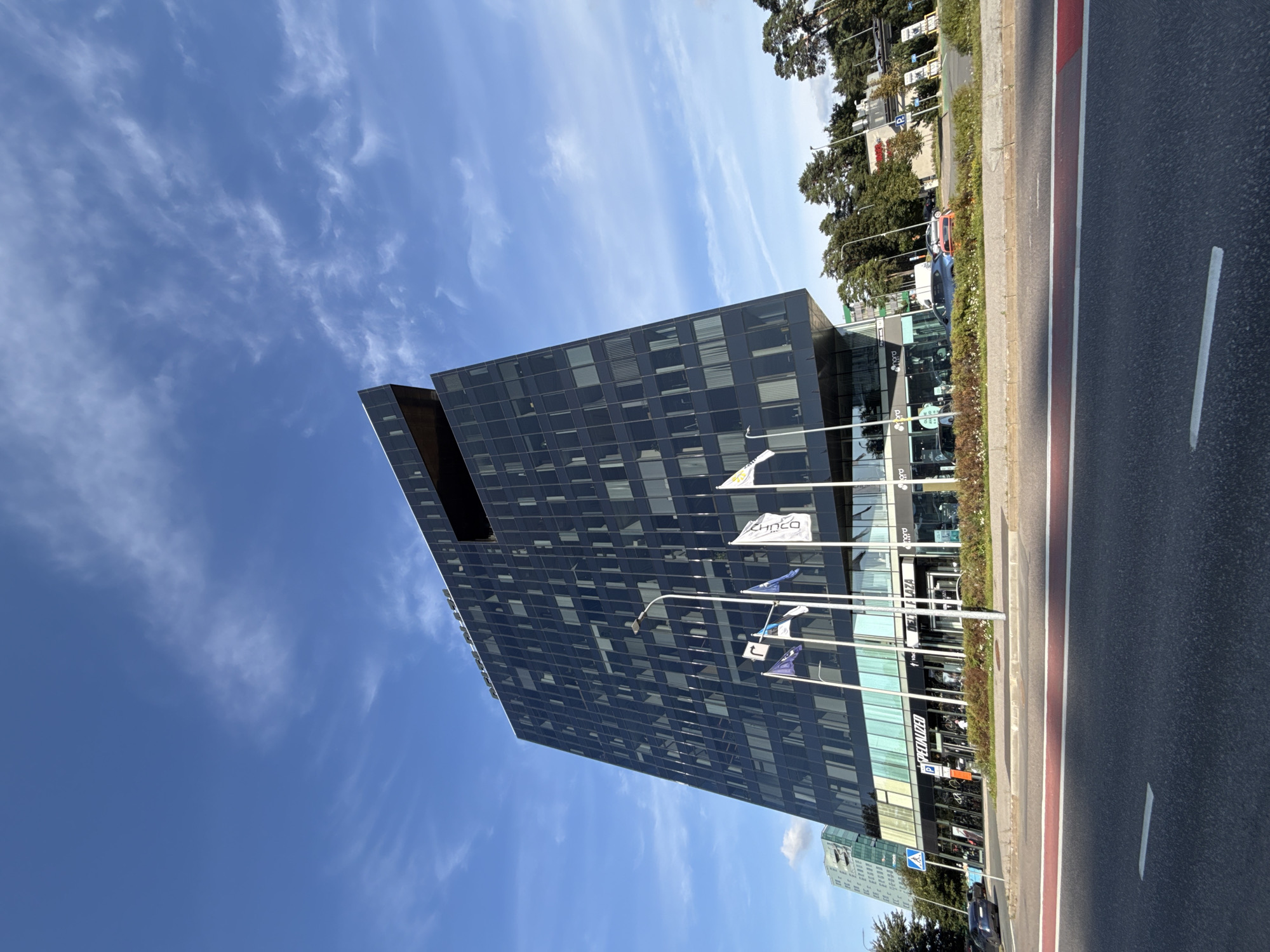 The image shows the modern architecture of the ADM Interactive building in Tallinn, Estonia, on a clear day with scattered clouds. The building, with its dark, reflective glass facade, is the central subject, angled from a low perspective, emphasizing its height and unique geometric design. In front of the building, several flags are planted on poles, including the Estonian flag. Businesses like "SPECIALIZED" and "NORD" are visible on the ground floor, suggesting a commercial and office space. A street with traffic lanes and a red bicycle lane runs in the foreground, with cars and a blue electric scooter parked nearby. Trees and other buildings, including a lighter-colored building in the background, are visible on the right side of the frame, indicating an urban environment. The sky is a vibrant blue with wispy white clouds.