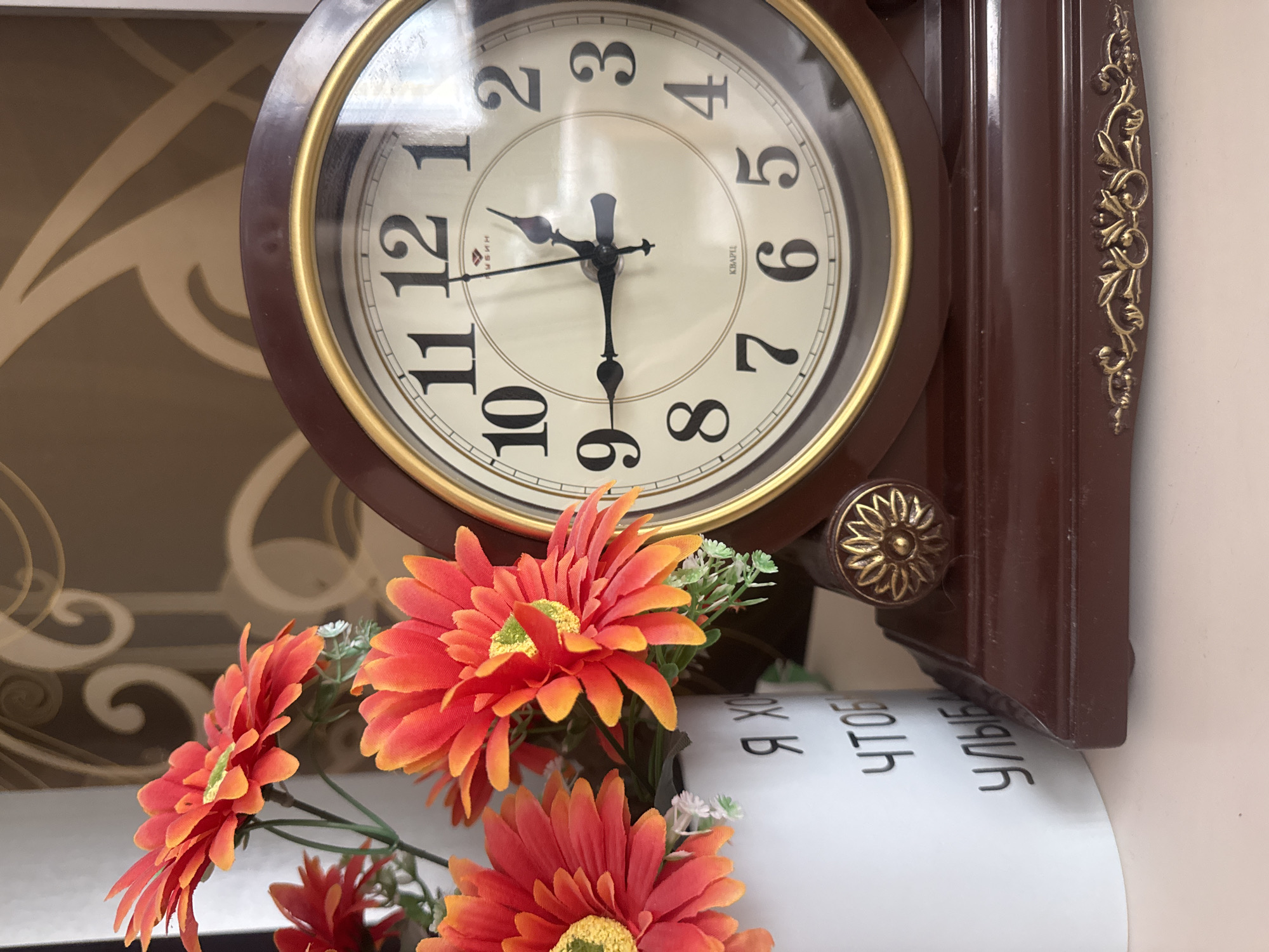 Here is a detailed description of the image:
The image features a wall clock positioned above a bouquet of artificial flowers. The clock is round with a white face and black numerals. The hour and minute hands indicate that the time is approximately 11:30. The clock has a brown frame with gold accents. Below the clock is a roll of white paper with writing in Cyrillic script. Arranged in front of the clock is a bouquet of orange and red artificial flowers. The background wall behind the clock and flowers appears to be beige. There is no visible indication of the city or specific location.