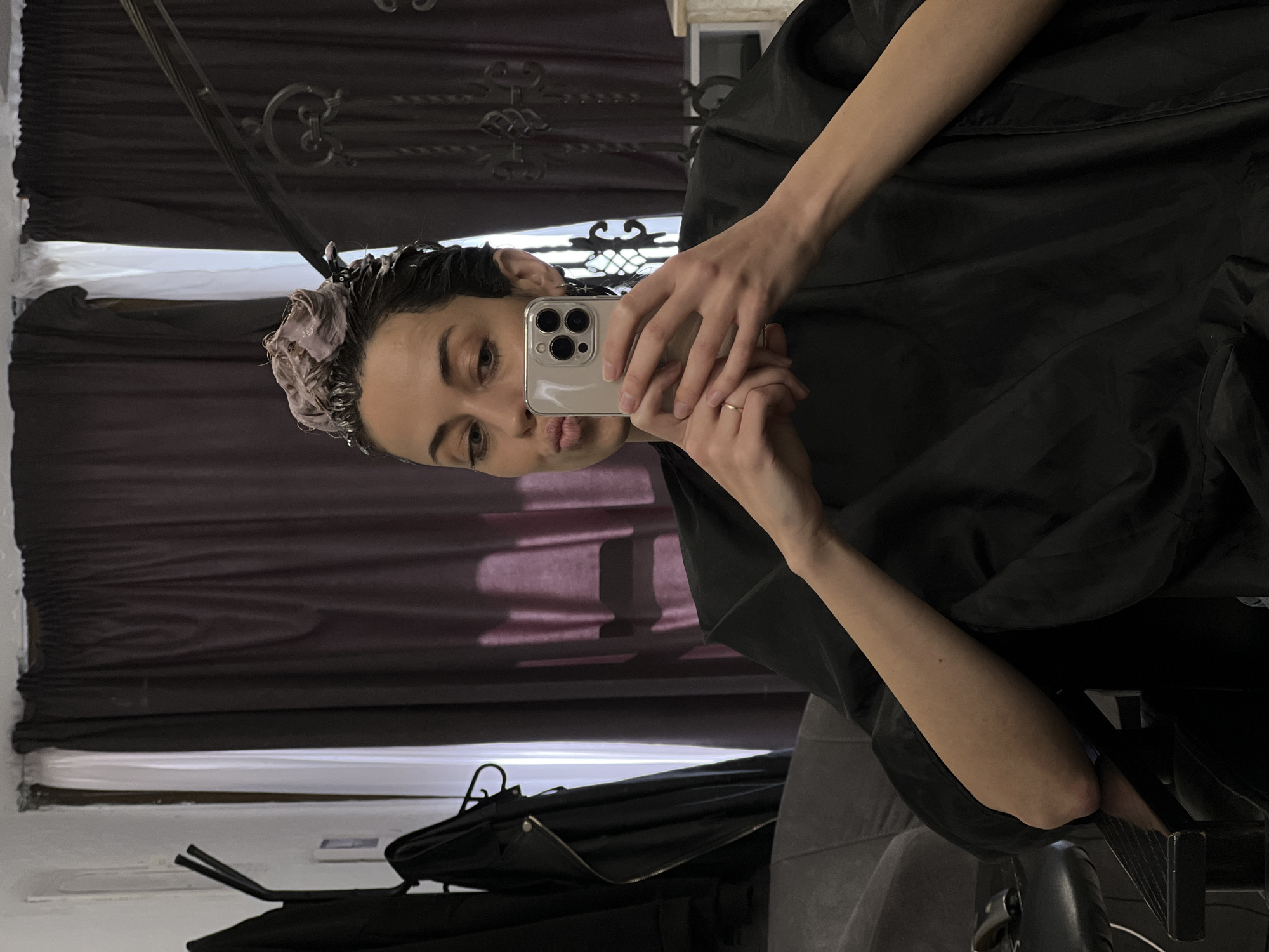 A person is taking a selfie in Bar, Montenegro. The person is wearing a black cape and has hair product in their hair. They are holding a phone to take the picture. The background includes a mirror reflecting a dark curtain and a window. A chair is visible in the lower portion of the image.