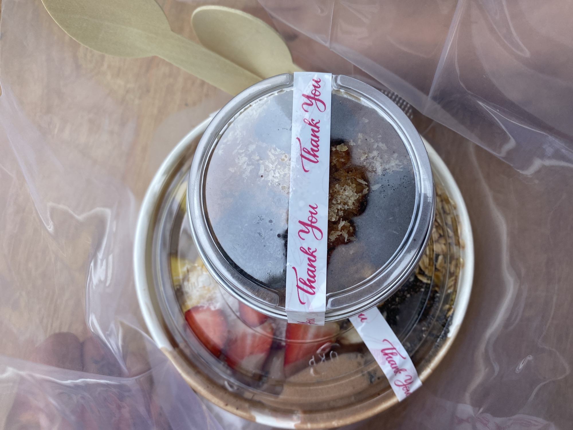 An overhead, close-up shot shows two take-out containers of food, one inside the other, and two wooden spoons, all within a clear plastic bag. The larger, outer container is a tan, paper bowl with a clear plastic lid. Inside this bowl, slices of fresh strawberries are visible on top of what appears to be yogurt and granola. The smaller, inner container is round and clear, with a dark, granular substance inside, topped with a few flecks of what looks like coconut or granola. A white paper band with pink cursive text that reads "Thank You" is wrapped around the lid of the inner container, with a portion of the same band also wrapped around the outer container's lid. The wooden spoons are light brown and are placed next to the containers within the plastic bag, with one spoon angled towards the upper right corner of the frame and the other resting below it. The background is a blurred wooden surface. The scene is set outdoors, with hints of a bright, possibly sunny environment visible through the plastic bag. The setting is in the city of Mui Ne, Vietnam, likely near a beach or resort given the casual nature of the food packaging and outdoor presentation.
