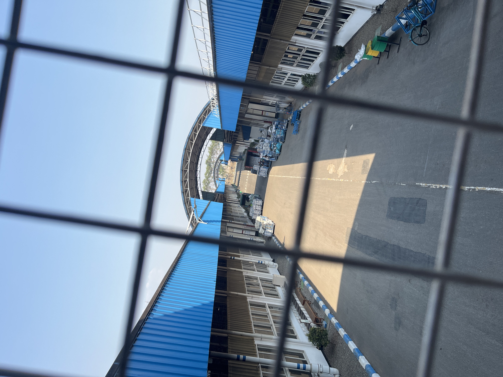The image captures an aerial view of an industrial complex, possibly a warehouse or factory, with long, parallel buildings featuring blue corrugated metal roofs. The complex is situated in Pelang, Indonesia, under a clear blue sky. A paved roadway runs between the buildings, with stacks of goods, some wrapped in plastic, visible on either side. Several carts and what appear to be trolleys are present on the roadway, suggesting ongoing activity. The buildings have numerous windows and large loading bay doors. In the foreground, a metal fence obstructs the view. The scene conveys a sense of industrial operation and logistics.