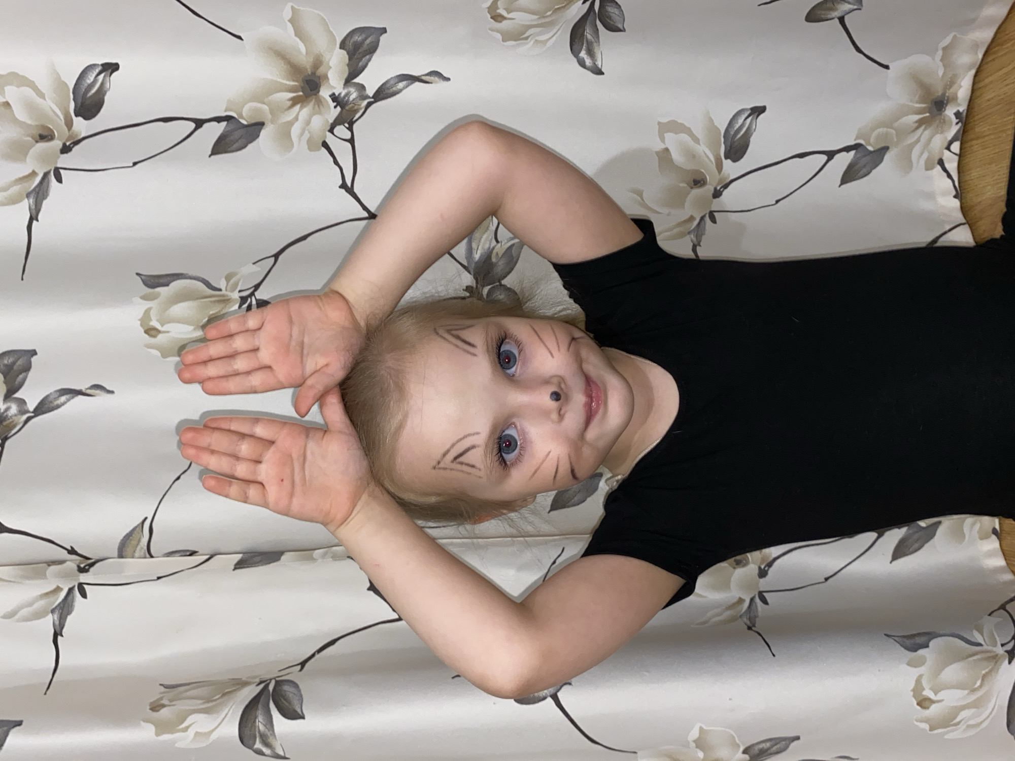 A young girl with light blonde hair and striking blue eyes is depicted lying on her back against a decorative, light-colored background featuring a pattern of muted white and grey flowers and branches. She is wearing a plain black short-sleeved top. Her head is positioned towards the right of the frame, and she looks directly at the camera with a gentle, playful smile.
Her face is adorned with cat-like makeup, including fine black lines on her cheeks resembling whiskers, and distinct V-shaped marks drawn above her eyebrows, evoking cat ears. A small black dot is visible on the tip of her nose. Further enhancing the cat costume, she holds her hands up, palms open, on either side of her head, mimicking the shape of cat ears. Her arms are bent at the elbows, framing her face.
The scene conveys a sense of innocent playfulness, with soft, even indoor lighting illuminating the child's joyful expression. No text is visible within the image. This photograph was taken in Votkinsk, Russia.