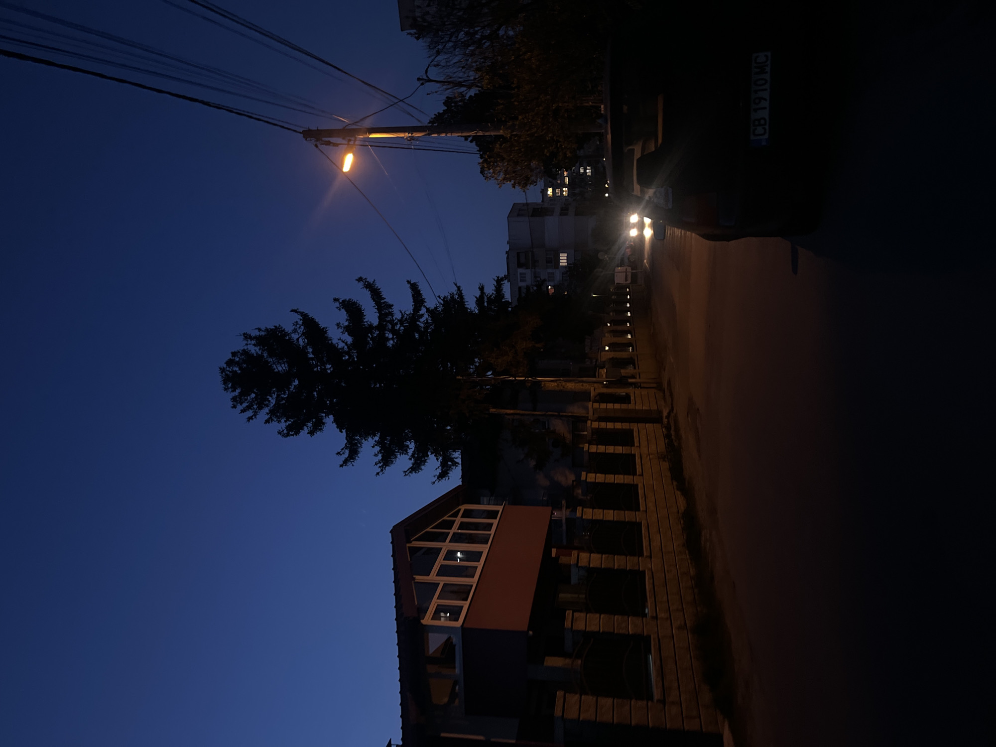 The image captures a street scene at night in Sofia, Bulgaria. The focus is on a residential area, with a building featuring a triangular window section prominently displayed on the left. A tall tree stands next to the building, silhouetted against the dark blue sky. A street lamp illuminates the scene from above, casting a warm glow on the surrounding area. Power lines stretch across the sky. On the right, a car with a visible license plate "CB 1910 MC" is parked along the street. In the distance, a multi-story apartment building can be seen with several illuminated windows. The street is dark. The image appears to be taken from ground level.