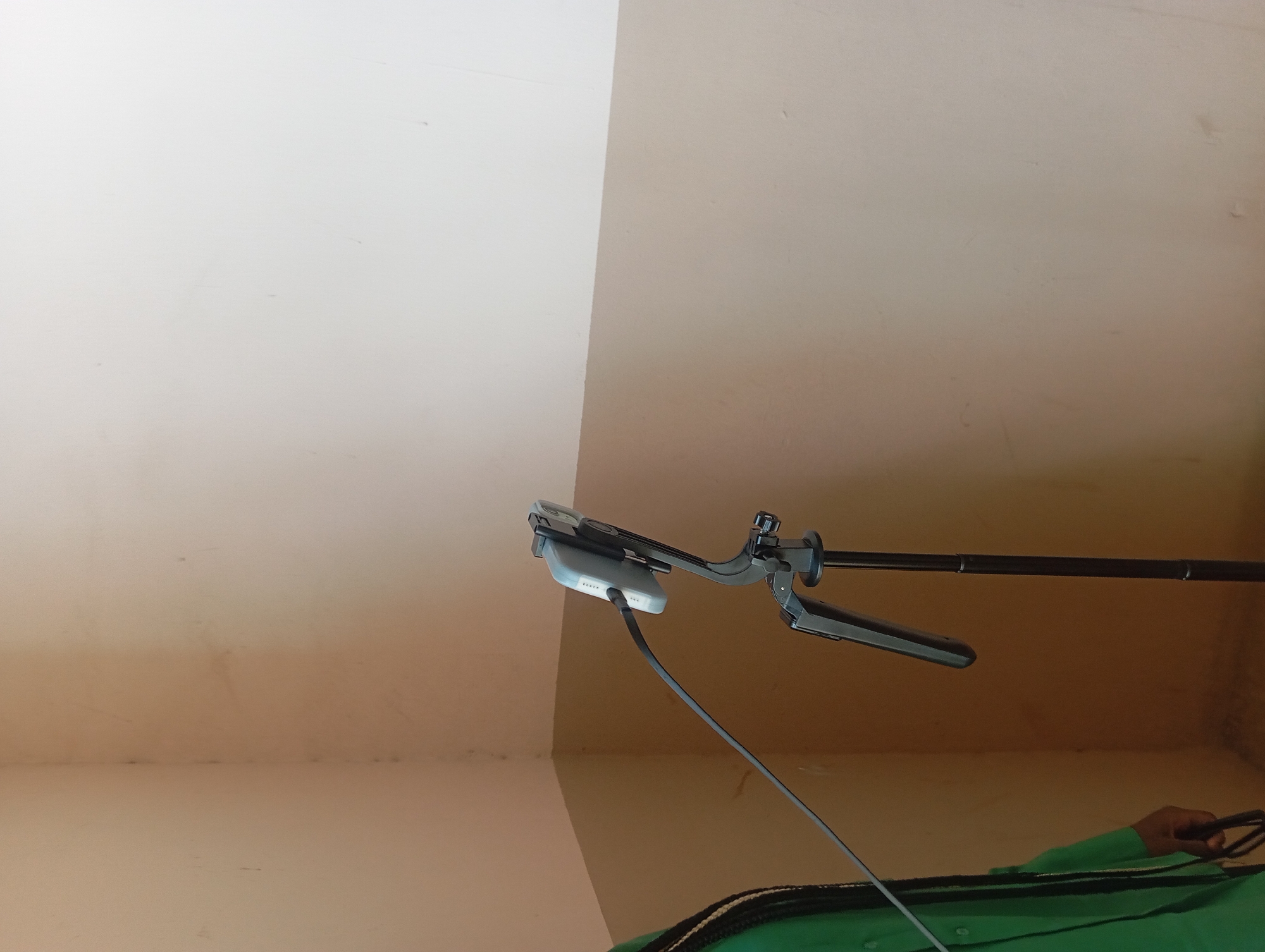In a room with beige and off-white walls, a person wearing a green shirt stands to the left of the frame. A black tripod is positioned in the center, supporting a phone holder with a light-colored phone. A dark cable extends from the phone to an unseen source. The lighting appears to be neutral.