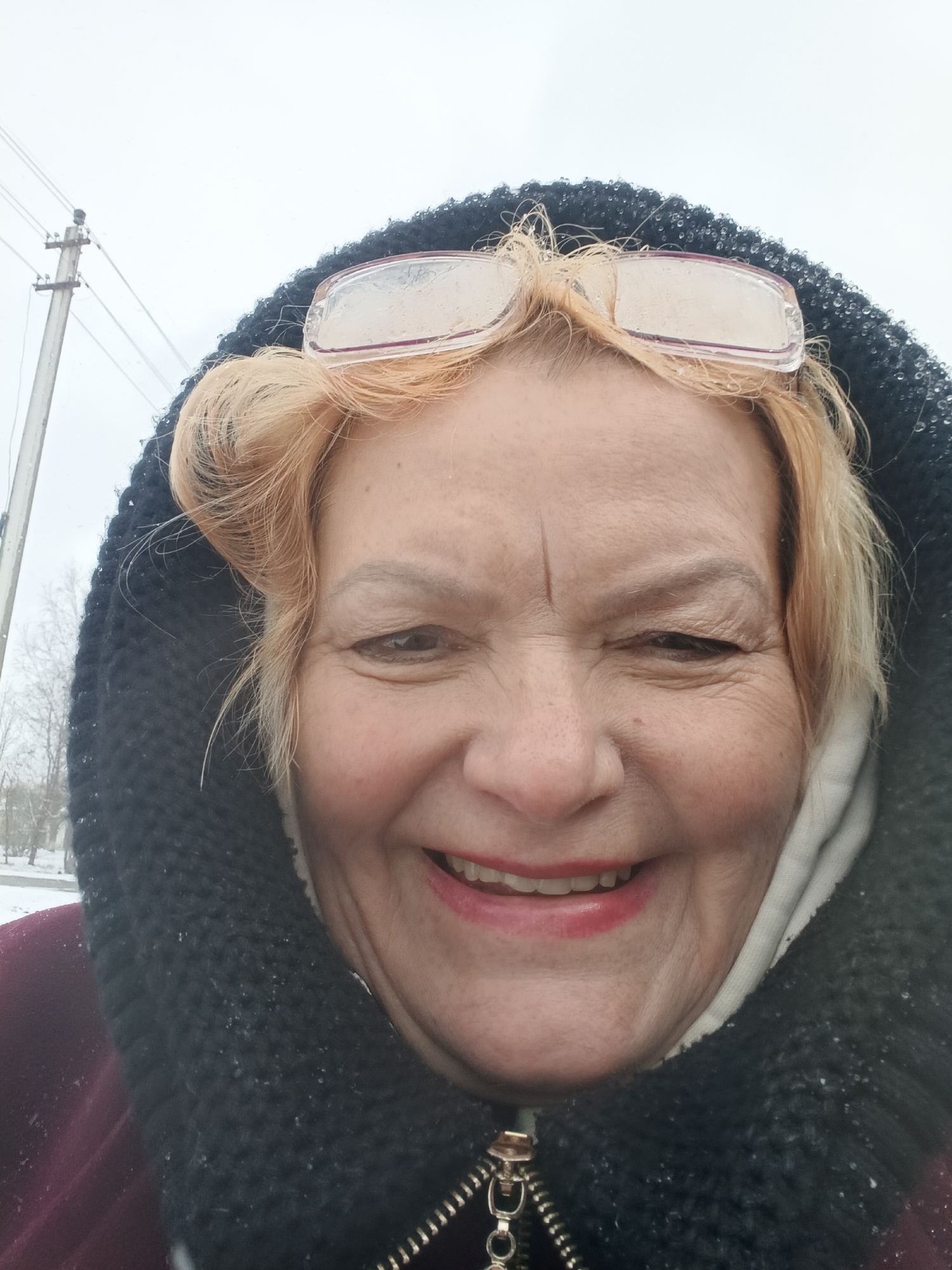 A close-up outdoor selfie captures an older woman with blonde or light red hair, fair skin, and a broad, joyful smile. Her eyes are crinkled at the corners, reflecting her happy expression. She is wearing a dark, possibly knitted, hooded garment that frames her face, and a burgundy or maroon jacket underneath, visible towards the bottom left. Clear-framed glasses are pushed up onto her forehead.
Small droplets or snowflakes are visible on her glasses and the textured fabric of her hood, indicating cold or inclement winter weather. The background consists primarily of a bright, overcast sky, suggesting daytime. To the left, a utility pole with wires stands against the muted sky, and sparse, bare trees are visible in the distance, further emphasizing a winter landscape. The ground also appears to be covered in snow. The woman's cheerful demeanor contrasts with the chilly environment of Красный Маяк, Russia. There is no visible text in the image.